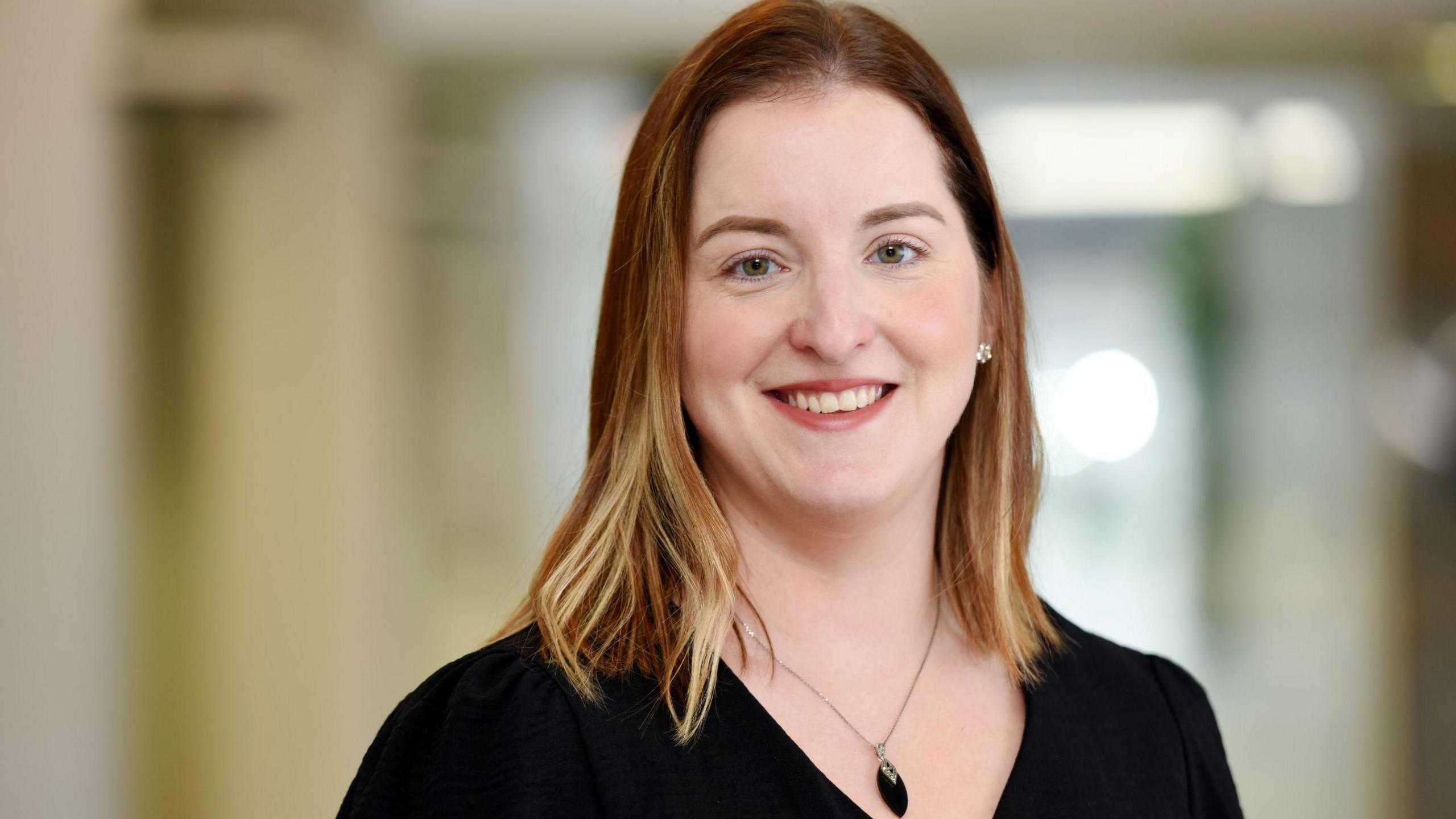 Professor Emma Giles, smiling at the camera. She has a shoulder-length blonde hair and is wearing a black top and a necklace with a black pendant. 