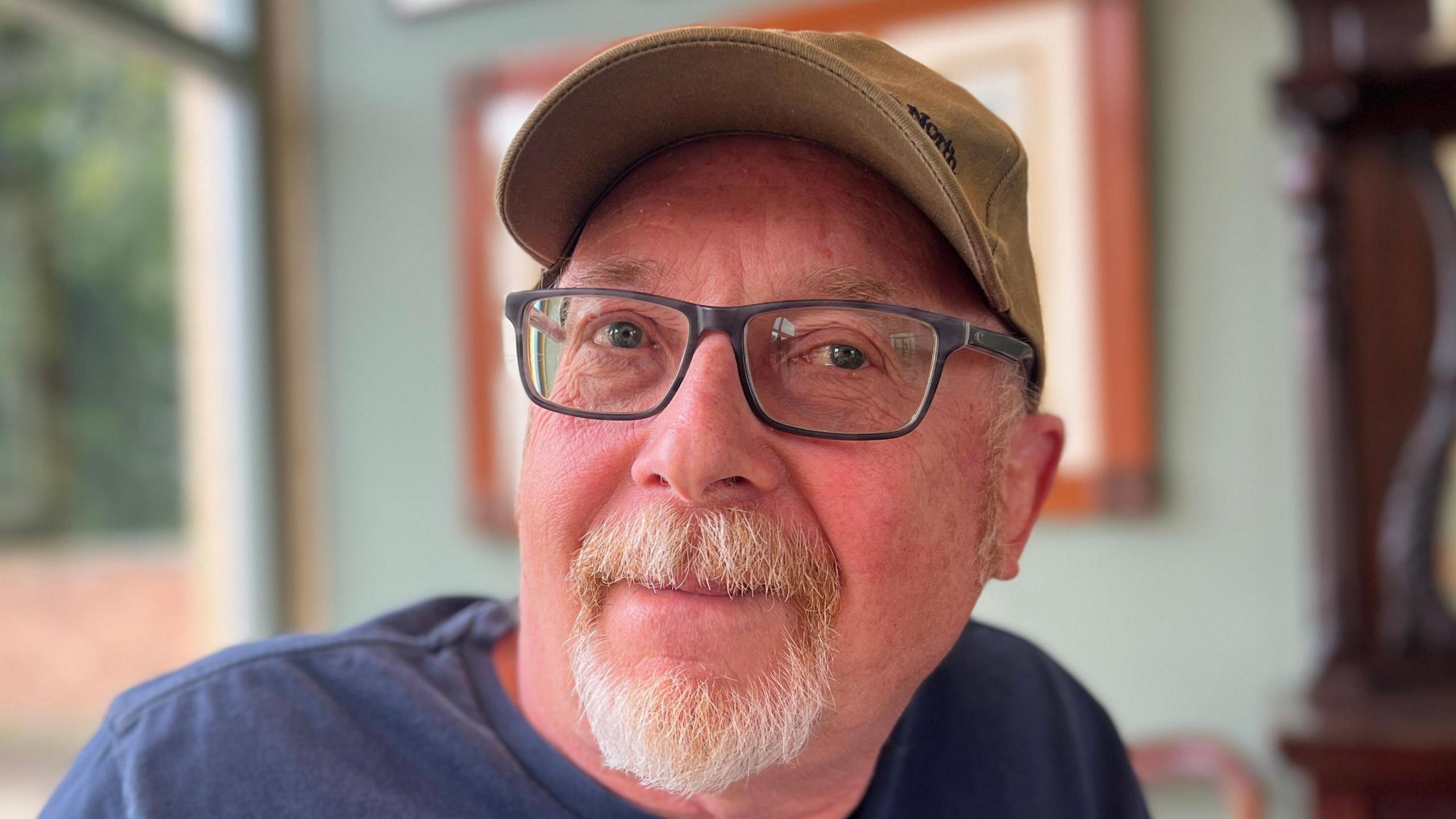 Colin Hopkirk is smiling, he has a ginger and grey moustache and beard. He's wearing dark blue glasses, a blue shirt and a beige cap.