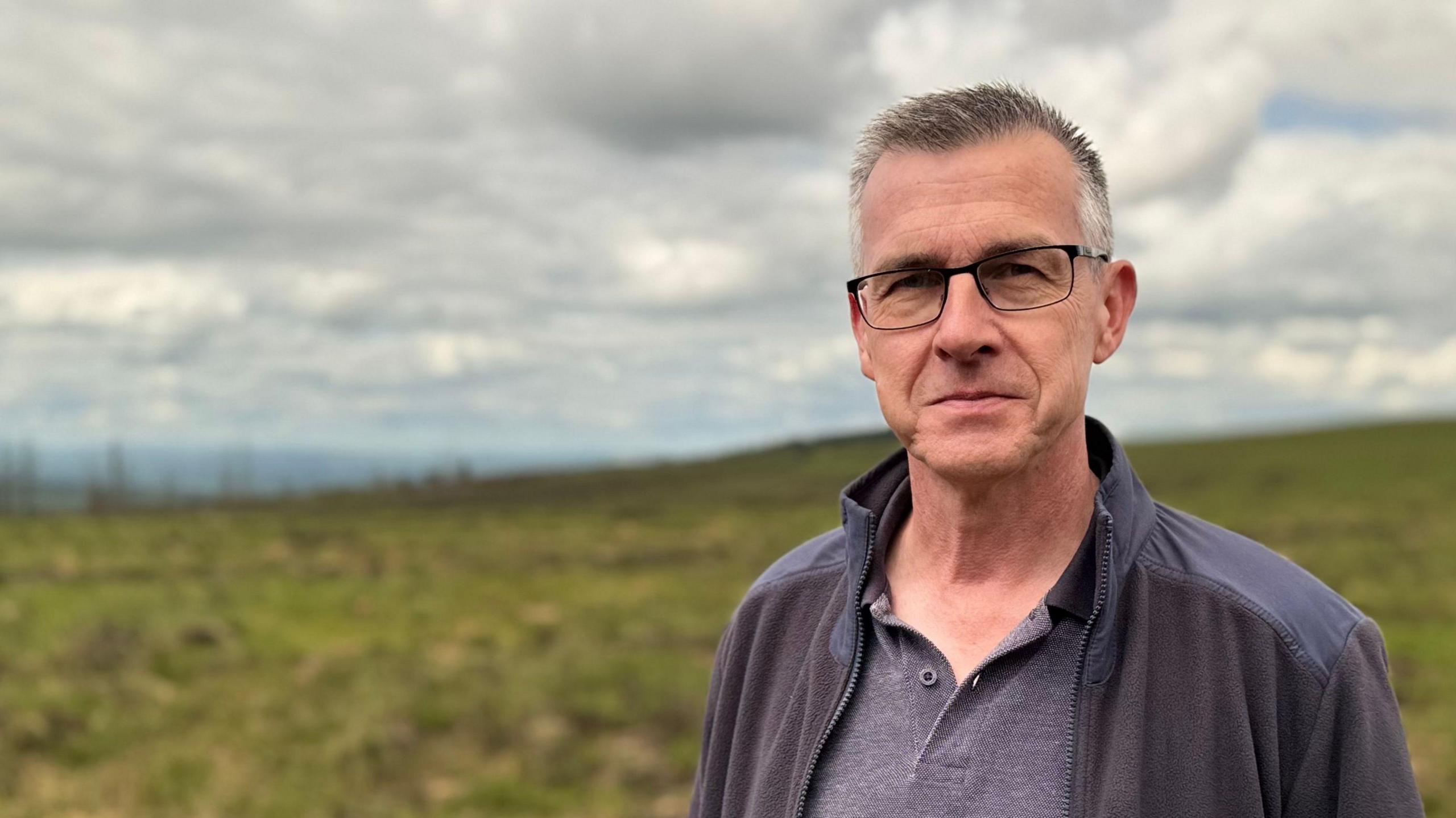 Mr McCabe has short grey and dark hair and is wearing rectangular glasss. He is smiling at the camera as he wears an unzipped fleece and a grey polo shirt. Behind him is the peatland which is mostly green.