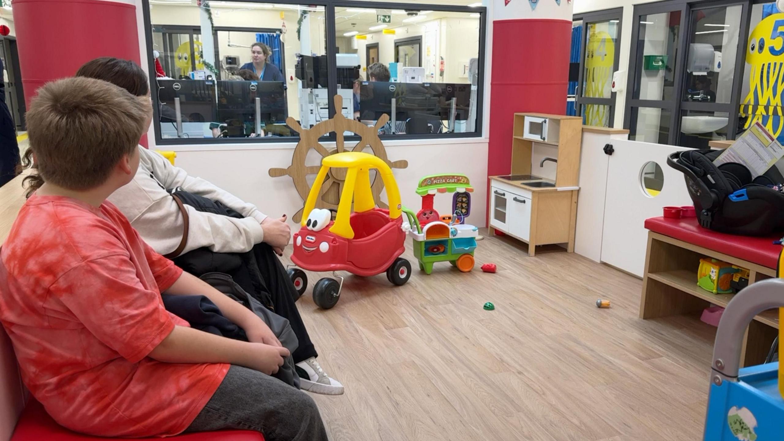 Children and parents sit in a modern play area with a play kitchen, shelves with toys. 
