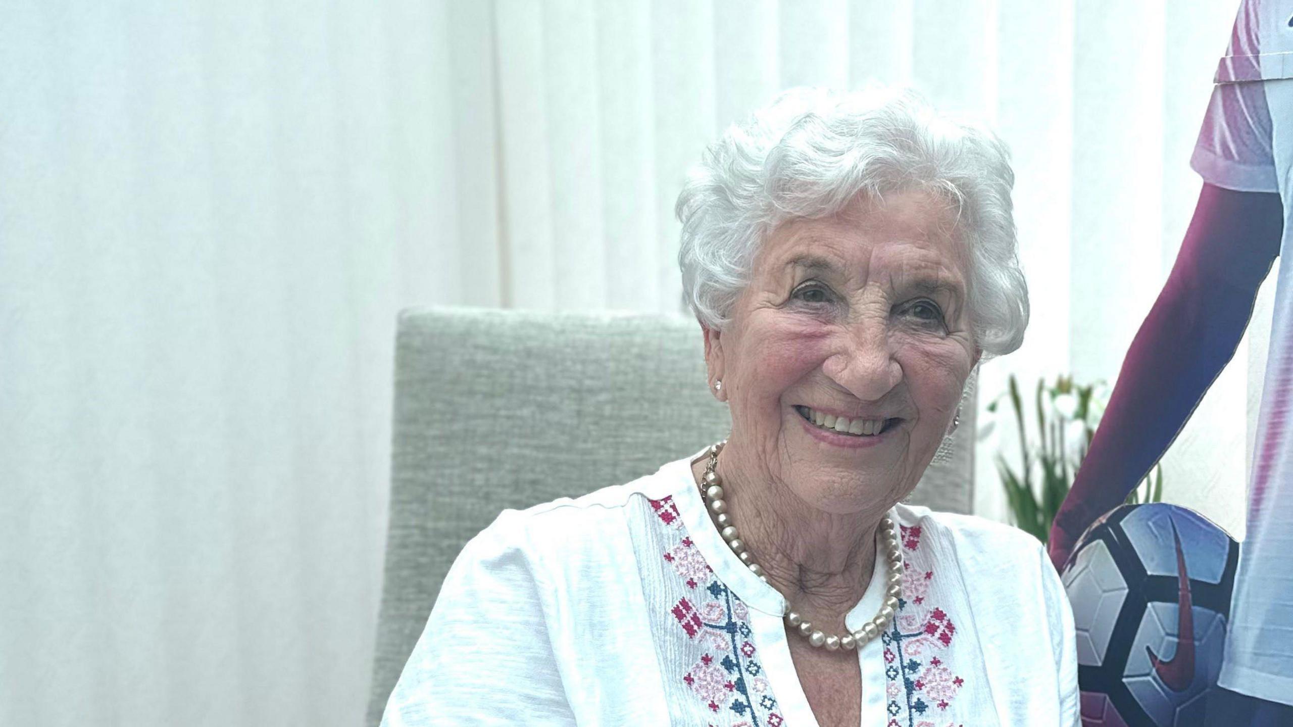 Gloria Stapylton sitting in a chair next to a life-size cardboard cut-out of her granddaughter Lucy Bronze. Mrs Stapleton is wearing a pearl necklace and a white blouse. She has short white hair and is smiling at the camera.
