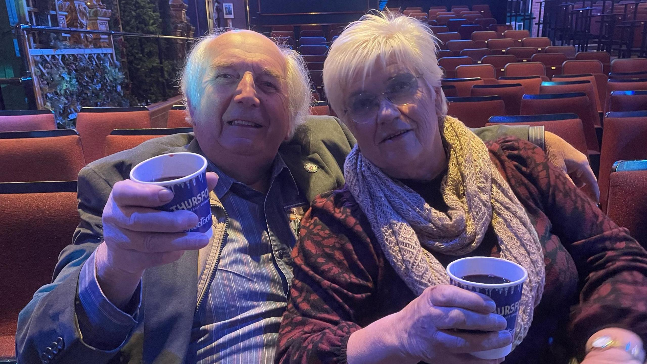 Brian and Joyce Cooper sitting on seats in the theatre each holding a drink in a paper cup. Brian is dressed in a grey jacket and a blue pinstripe shirt. Joyce is wearing a red and black patterned outfit with a cream coloured scarf, and she has round glasses. They are both looking at the camera and smiling.