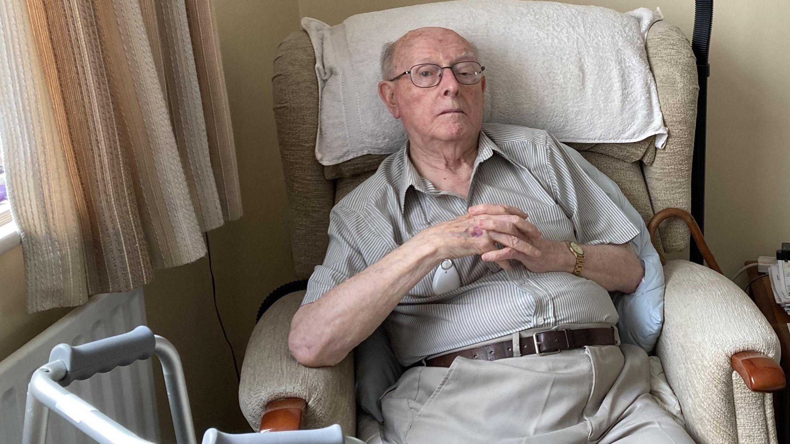 Garrod Evans sitting in a chair with his hands folded, with a walking frame and walking stick nearby