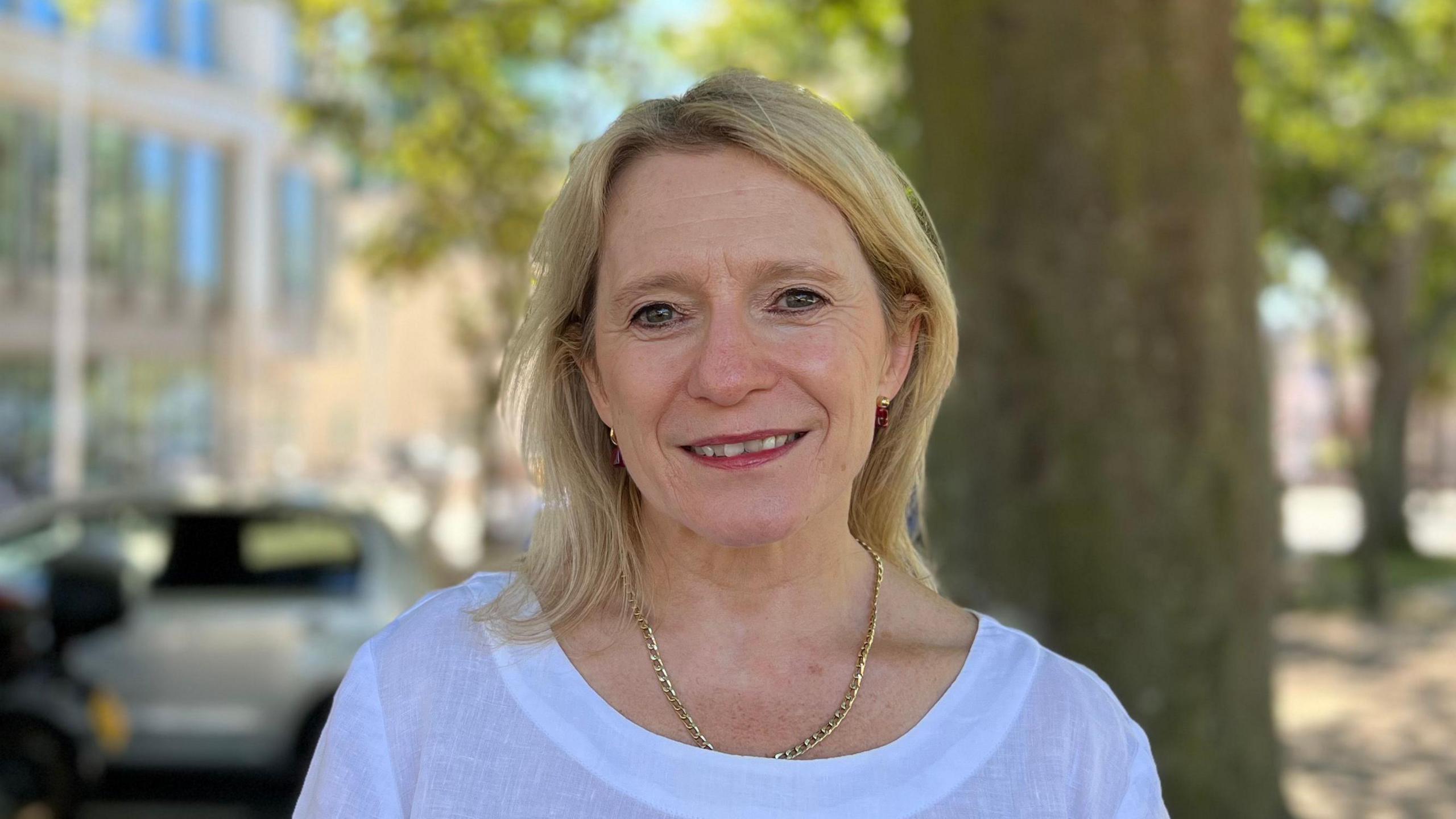 A person wearing a white top stands outdoors beside a large tree on a sunny day. Office buildings, parked vehicles and leafy green branches appear softly blurred in the background.