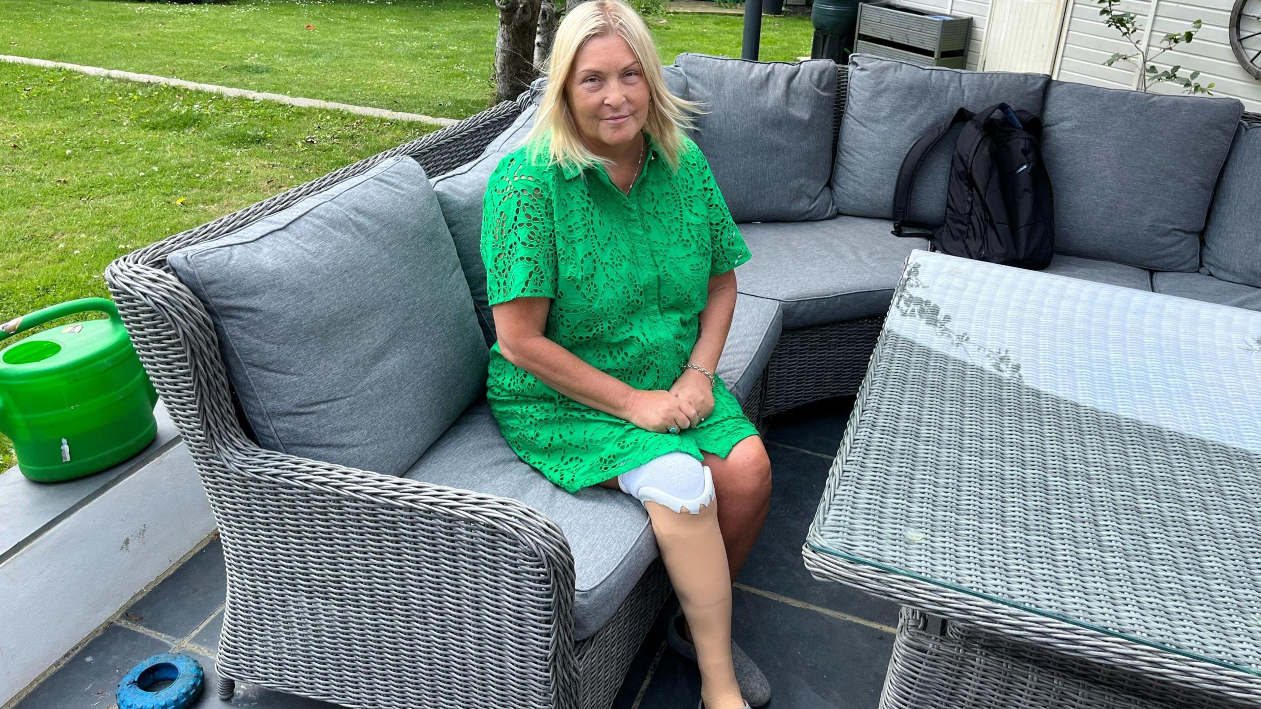 A woman with blonde hair and a green top sits on a grey corner sofa showing her prosthetic leg.