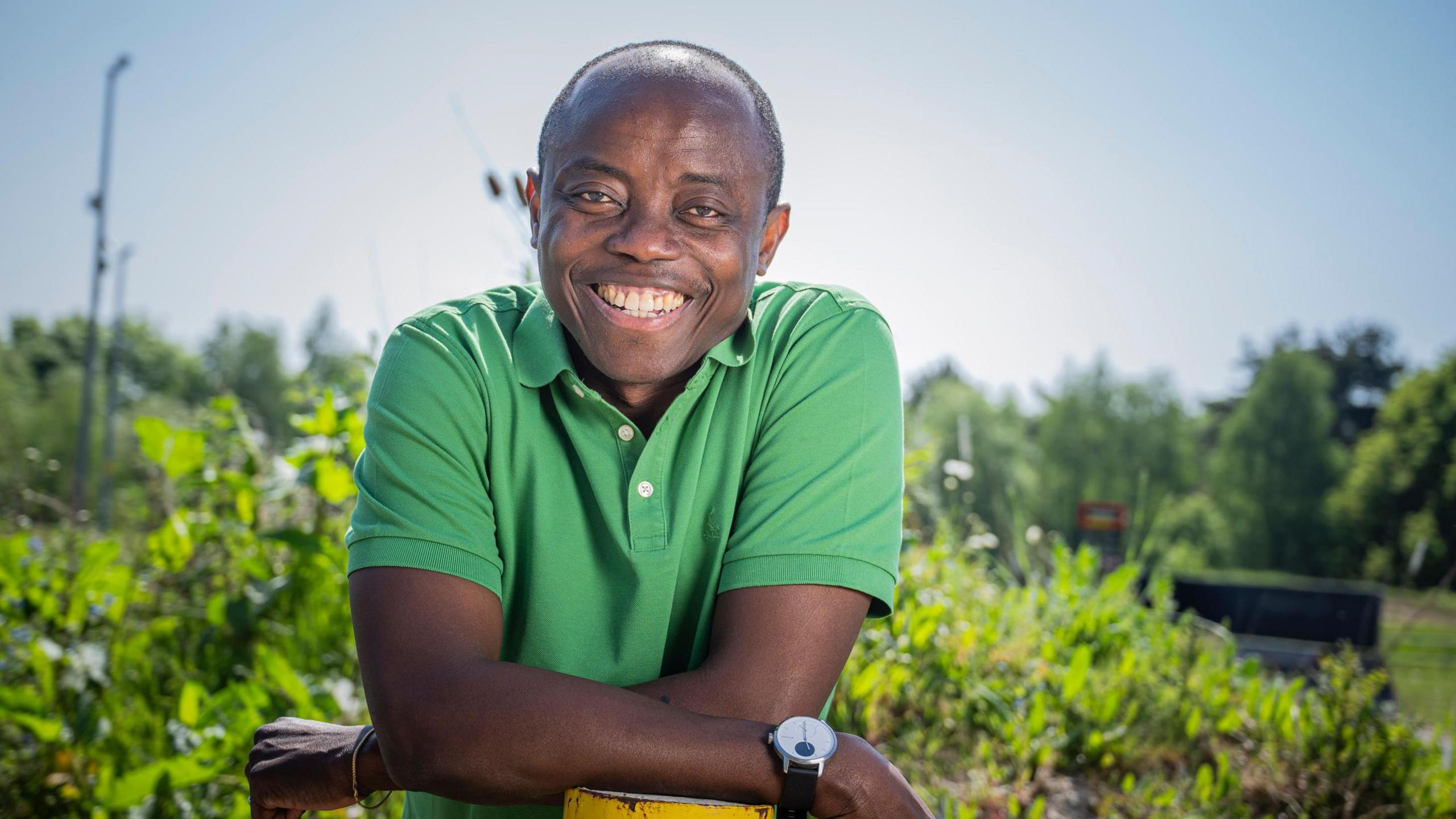 Abiola smiles broadly. His hair is shaved back and he wears a green short-sleeved shirt. He is resting his elbows on a post in a garden setting.