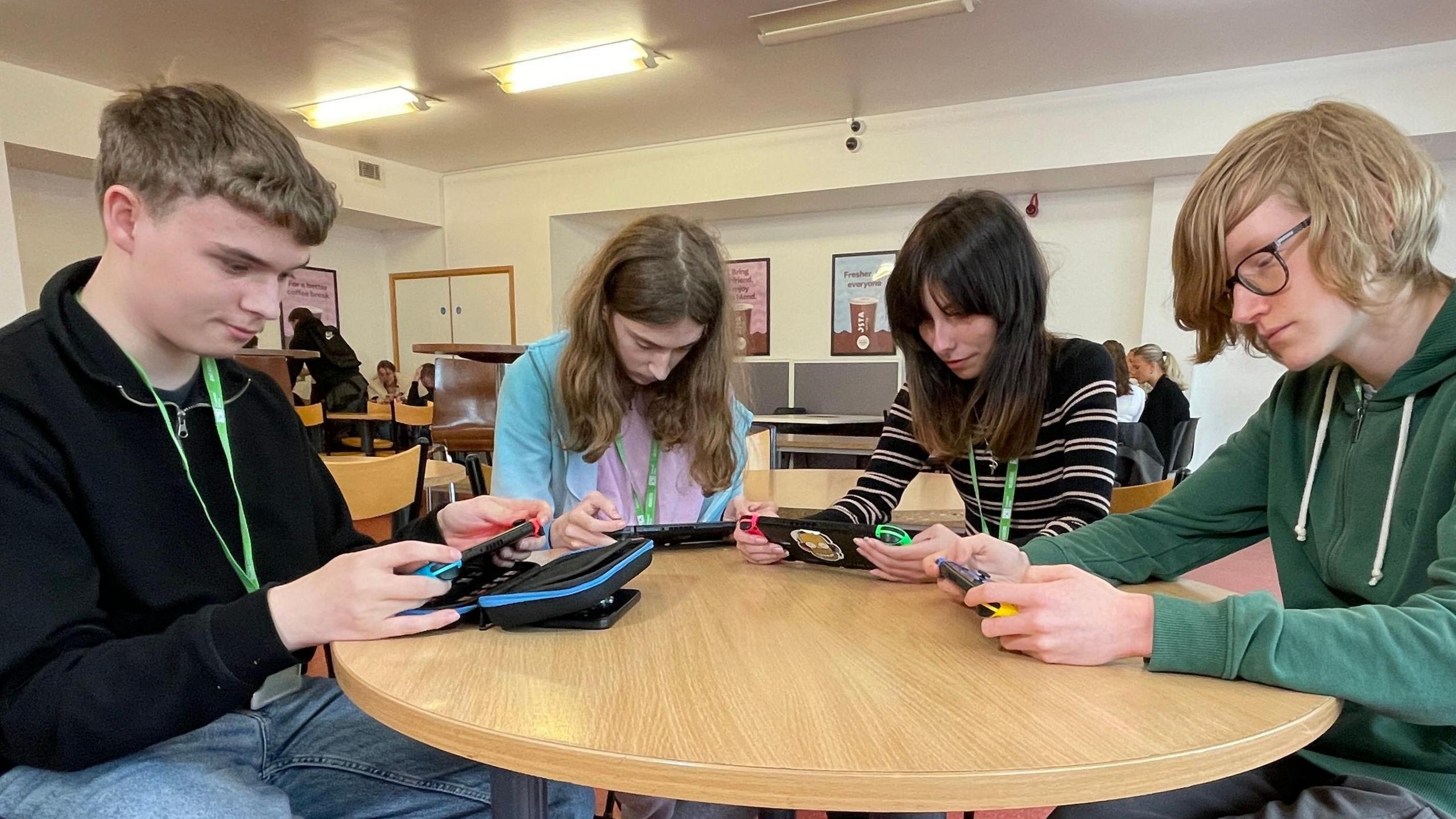 Four students are sitting at a wooden round table at the college coffee shop, each looking down playing a different game on their Switch consoles.