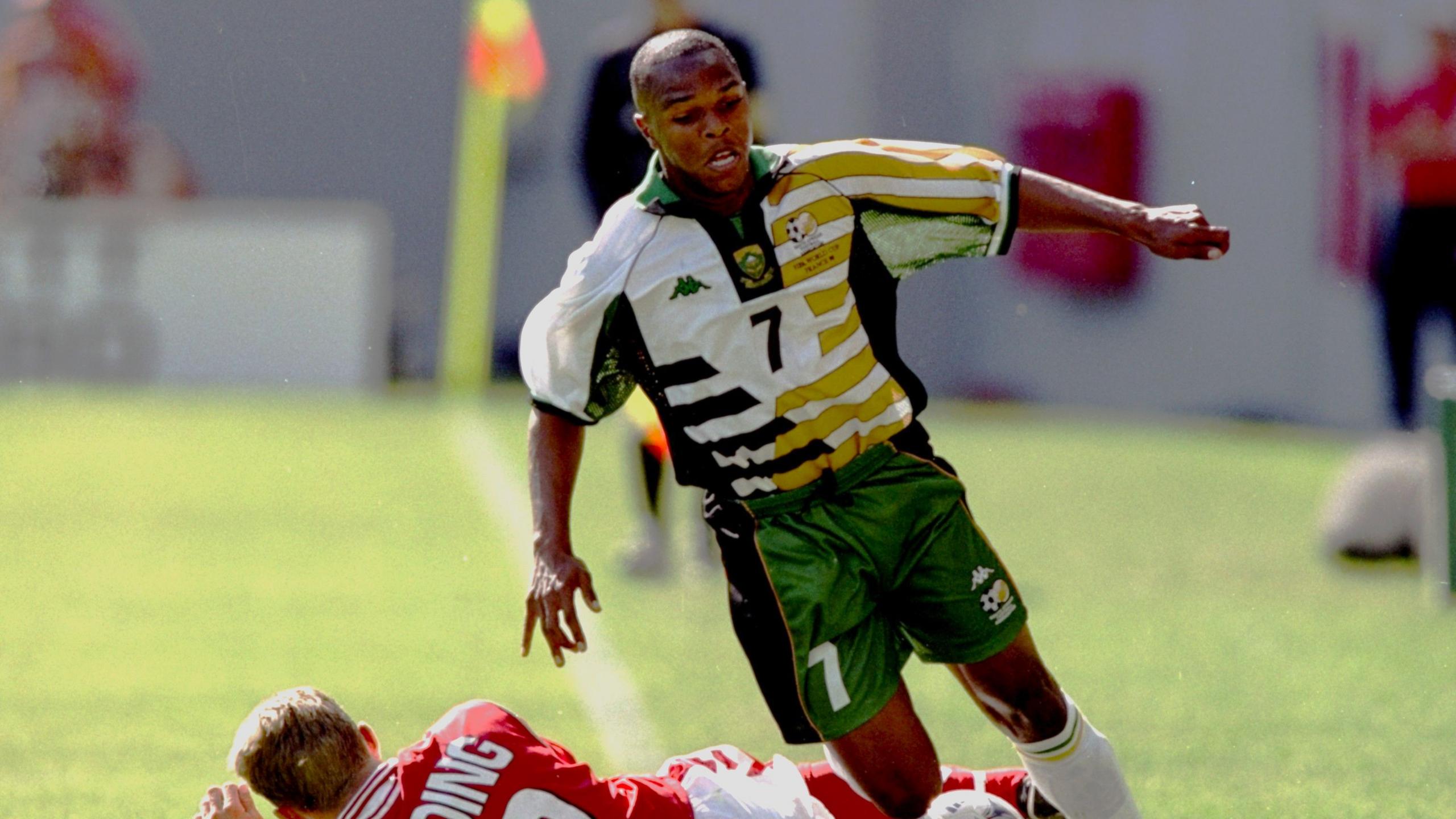 An image from the 1998 World Cup sees South Africa's Quinton Fortune running with the ball down the touchline, looking down towards the ball as he leans to his right to keep it in play. A Danish defender in a red shirt can be seen sliding across the turf behind Fortune, who has just evaded the tackle. Fortune wears white socks, green shorts and a white, gold and green jersey which is made up of many oblong blocks positioned horizontally, with green ones on the left and gold on the right. In the centre of his chest is a green number seven