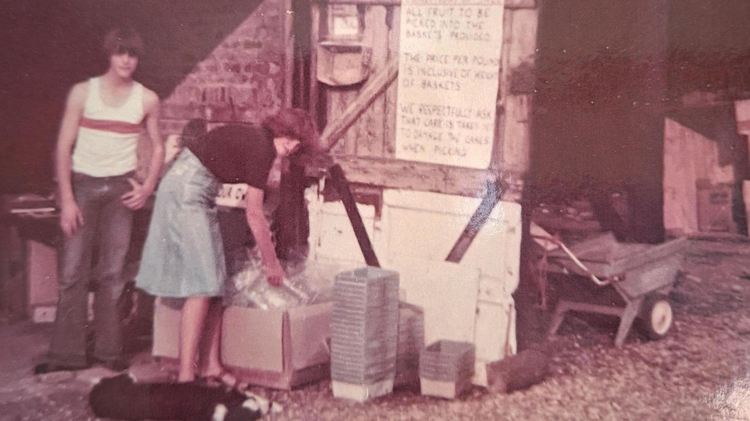 Colour photo from 1978 show Paddy Ivens in jeans and white cropped vest top with his sister, Karen who is bending over to get some plastic trays out of a cardboard box. She is wearing a denim skirt and dark short sleeved top, they are standing in what looks like the doorway to a barn or outbuilding.