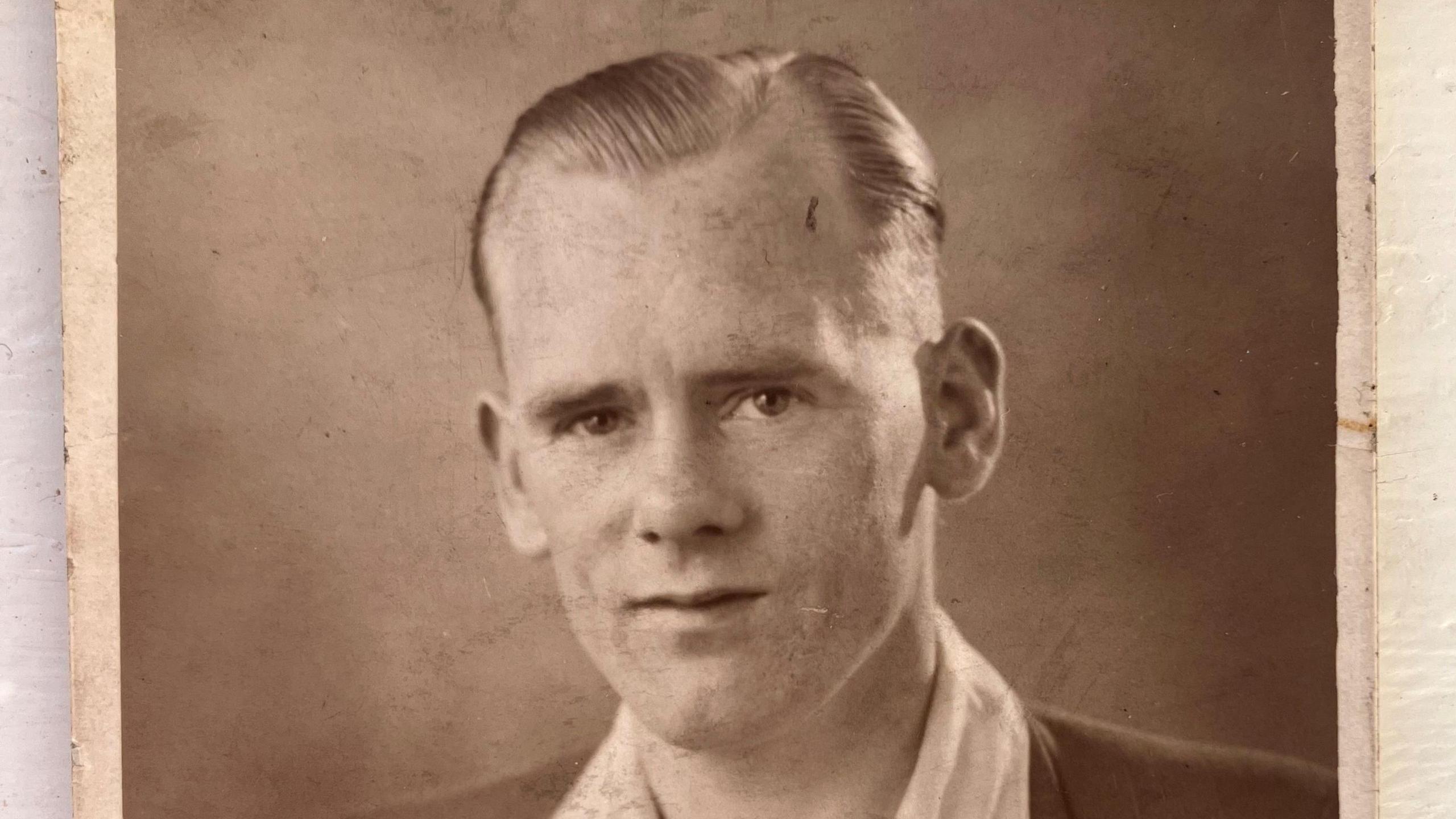 A sepia-toned portrait photograph shows an individual dressed in a formal suit jacket with wide lapels over a light-colored collared shirt. The jacket has a small pin on the lapel and a pocket on the left side. The photo has worn edges, indicating age, and is placed on a light surface.
