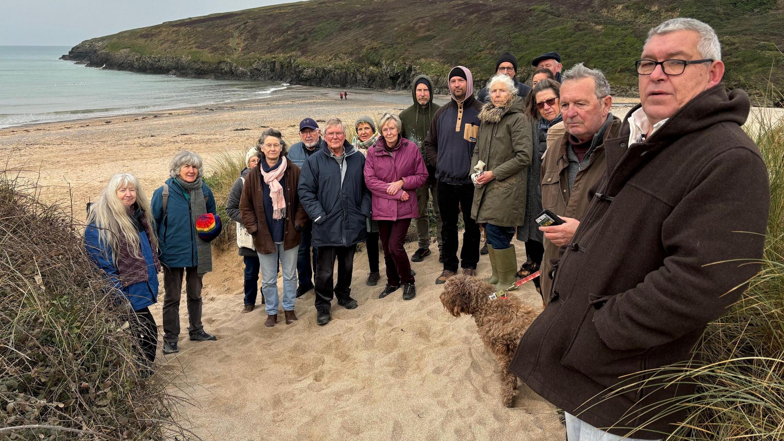 About 15 people standing on the sandy beach at Porthcothan Bay. They are bundled up in coats, scarfs and hats. There is a brown shaggy haired dog on a lead in the foreground.