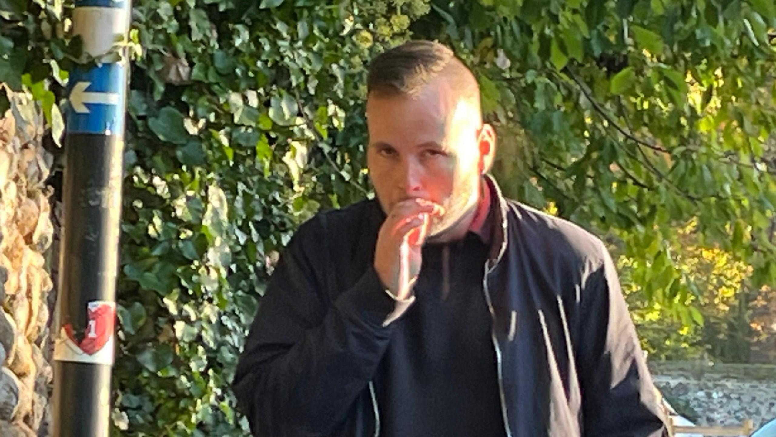 Wayne Finney arriving at Norwich Crown Court. His hair is shaved at the sides and long on top. He is wearing a black top with a black jacket on top and is smoking a cigarette. 