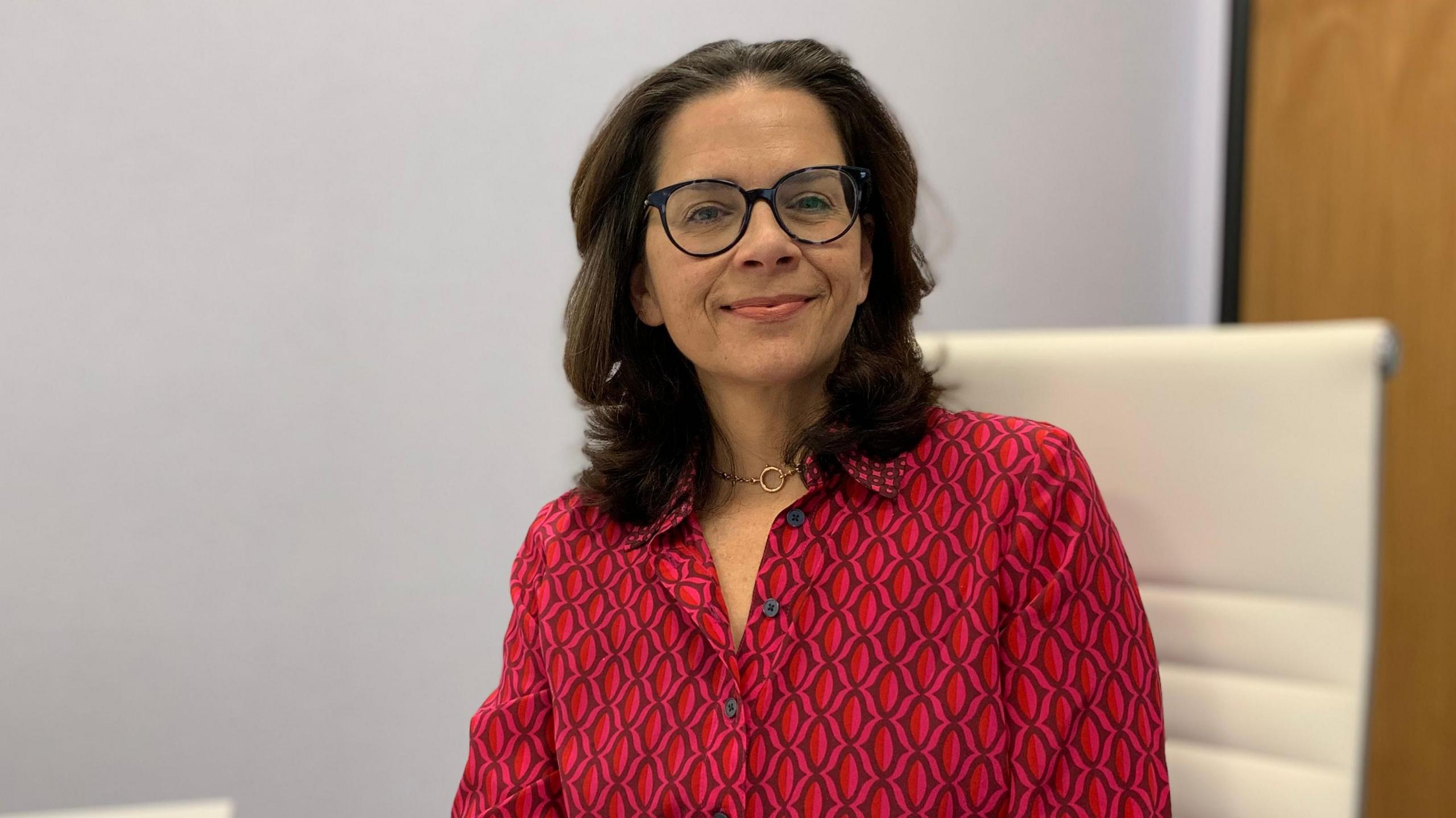 A lady with medium length brown hair and black rimmed glasses smiling at the camera. She is wearing a red and black shirt. 