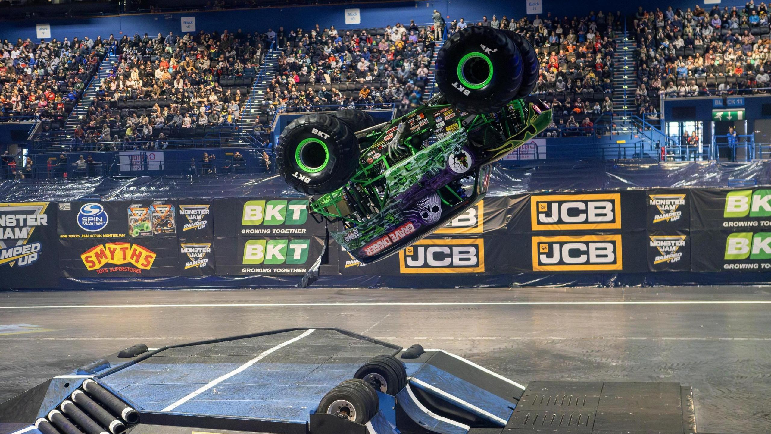 A monster truck mid-way through a flip in the air, above a large ramp in an arena. There are hundreds of people in the stand behind it. The truck is green and purple and has a skull on the side, as well as the words "grave digger"