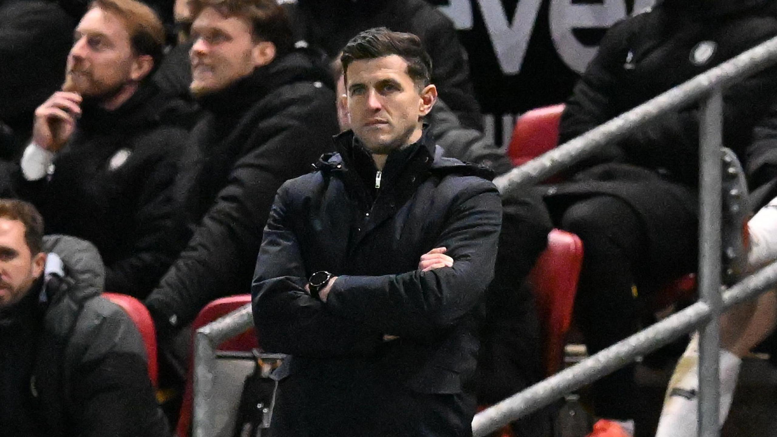John Mousinho standing on the side line of the football pitch with his arms crossed, and looking disappointed.