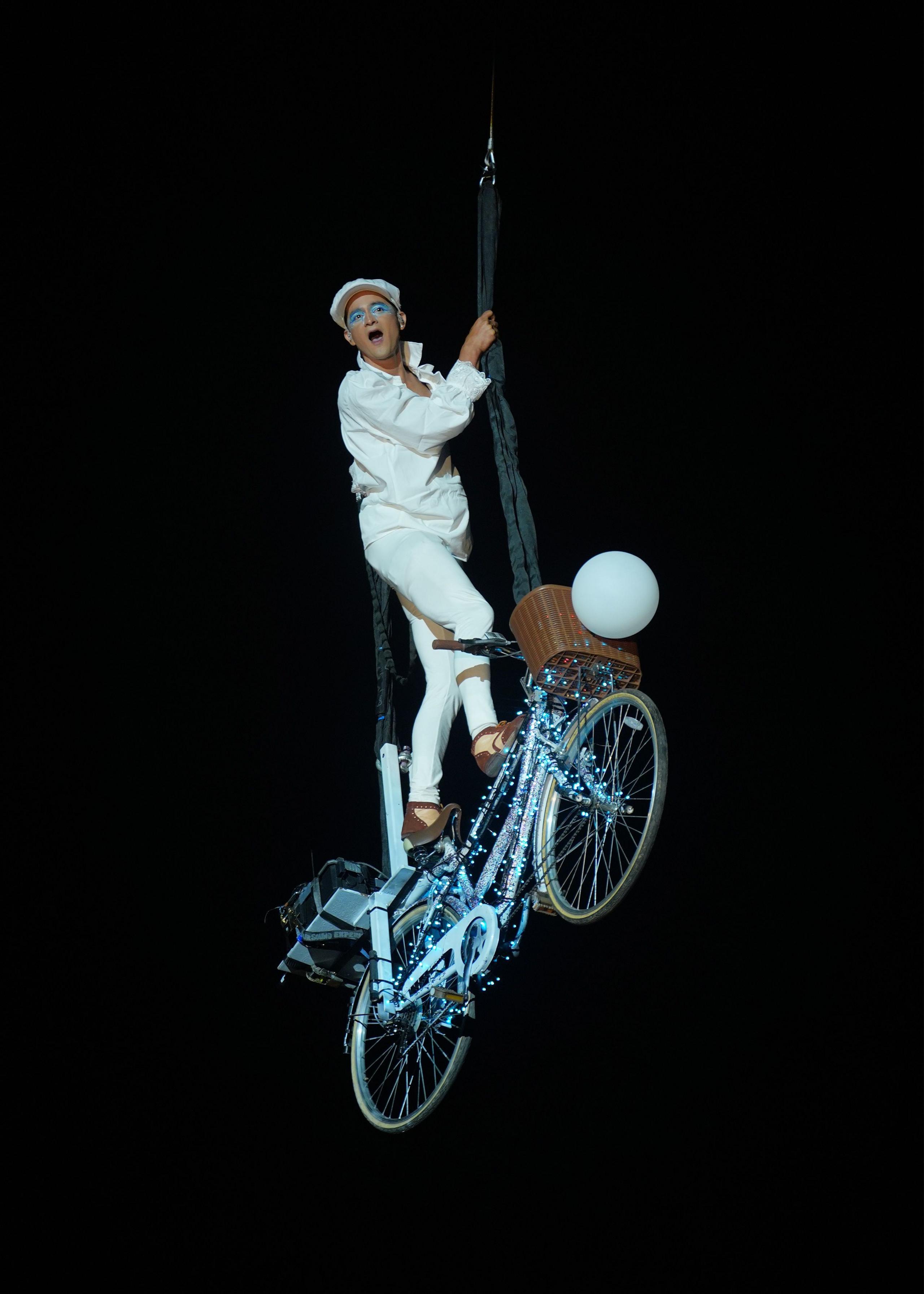 A man in white clothing stands on a lit-up bike, which is suspended in the air.