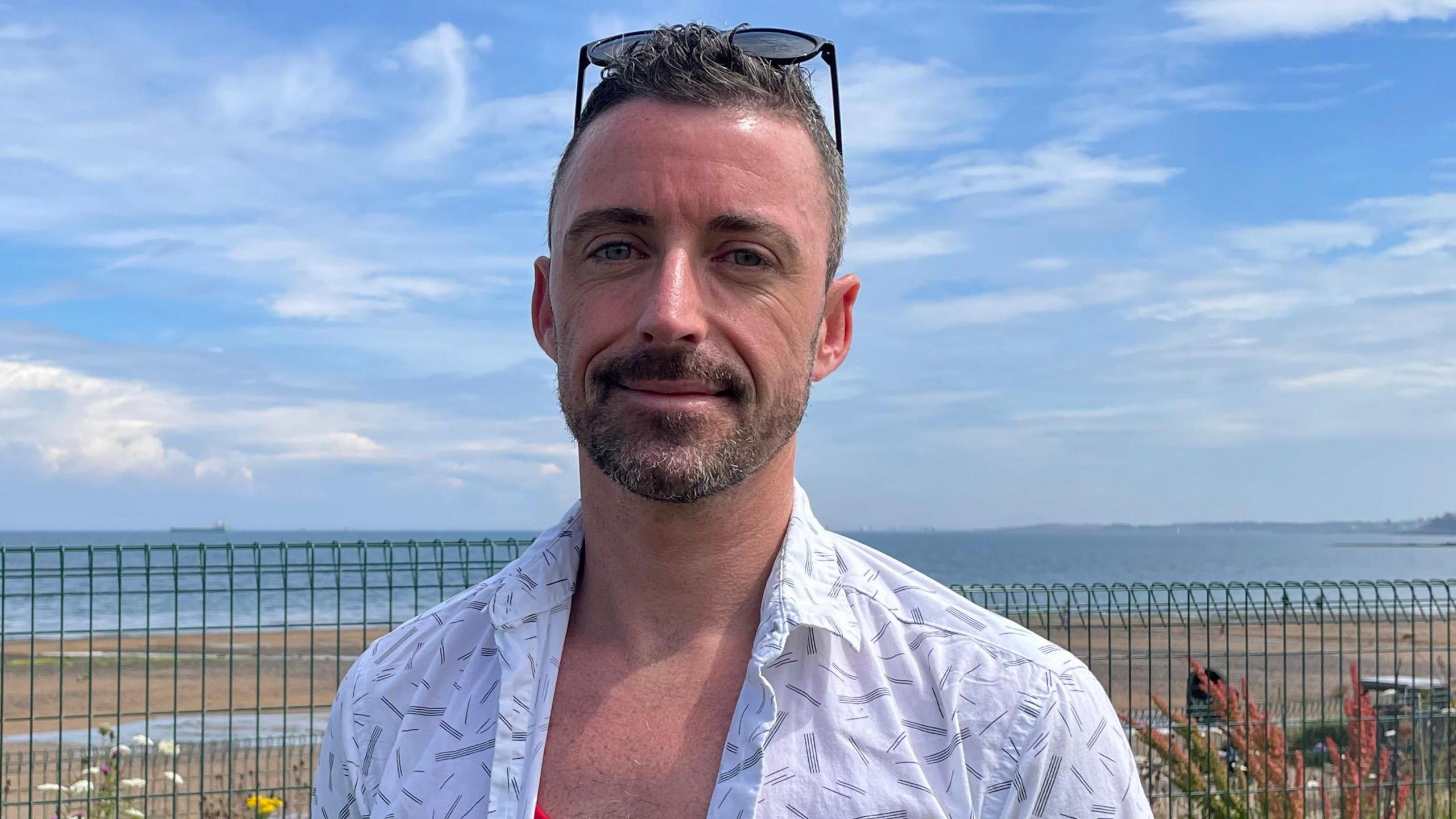 A man with short brown hair, wearing a red top and a white short sleeved shirt over it. He is standing in front of a beach. 