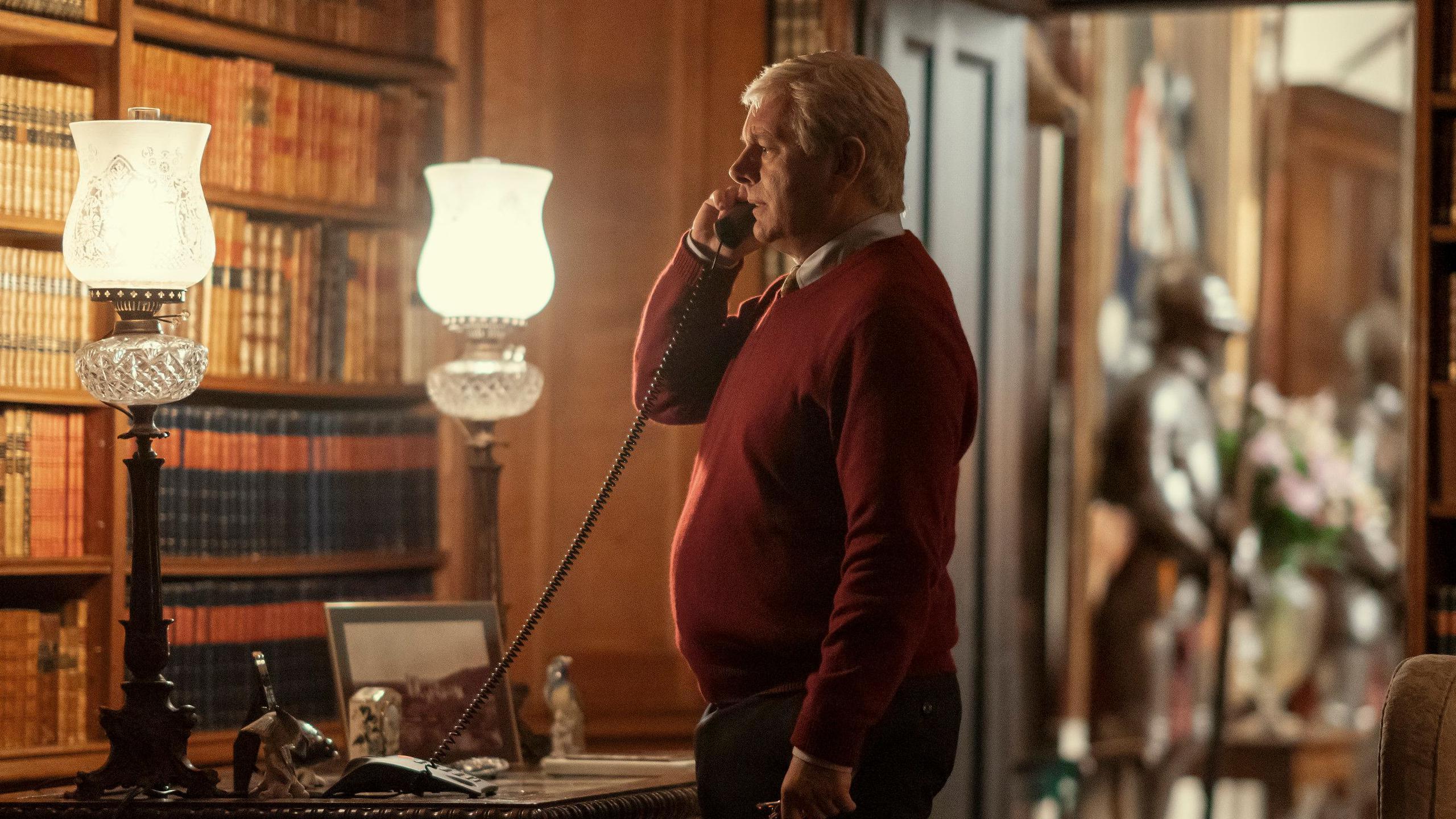 Michael Sheen, playing Prince Andrew, in a red jumper stands next to a desk, holding a telephone to his ear.