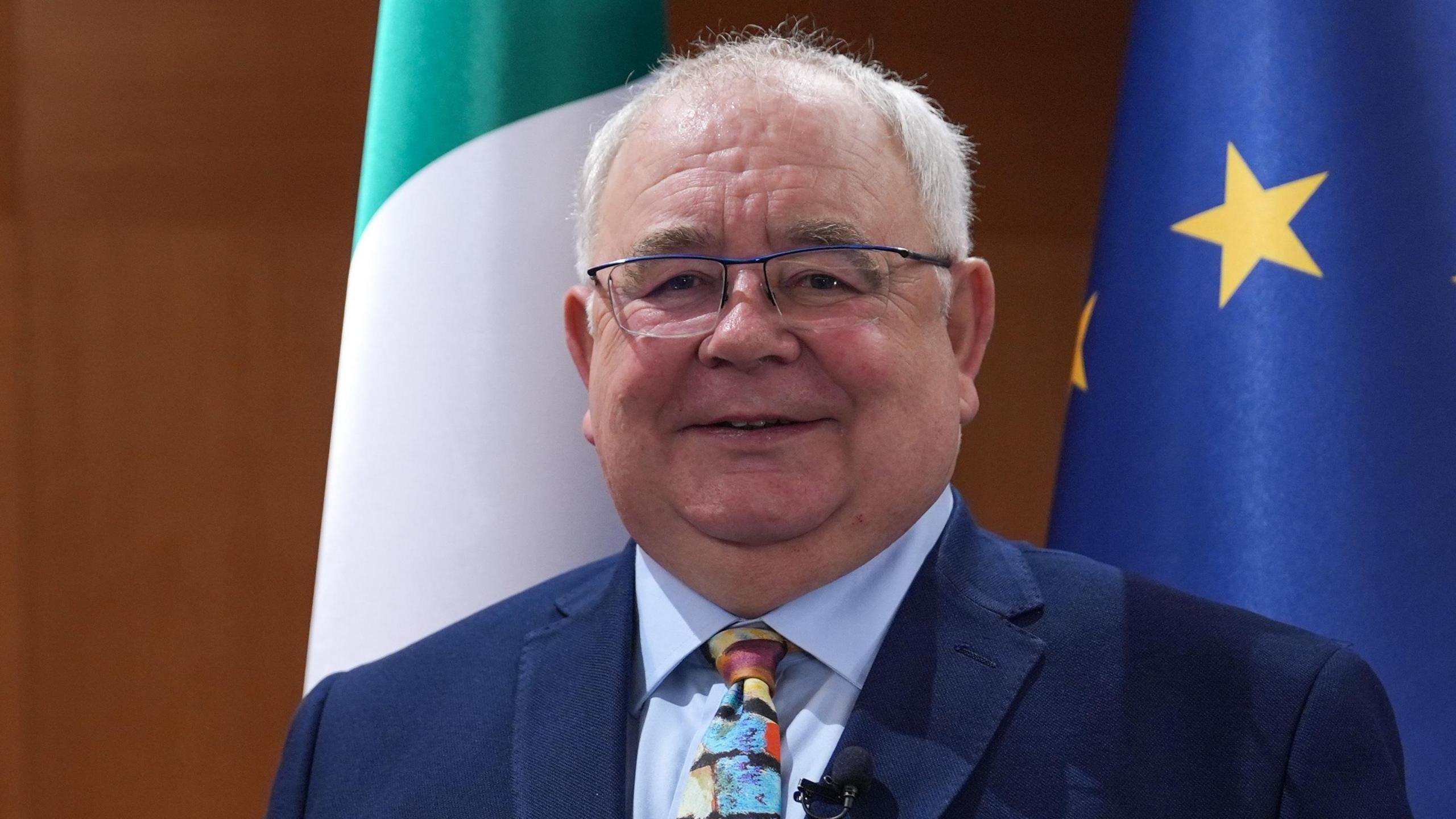 Sean O Fearghail is standing in front of the Irish tricolour flag and the Eu flag. He wears glasses and is wearing a navy suit with a blue shirt and a printed tie.