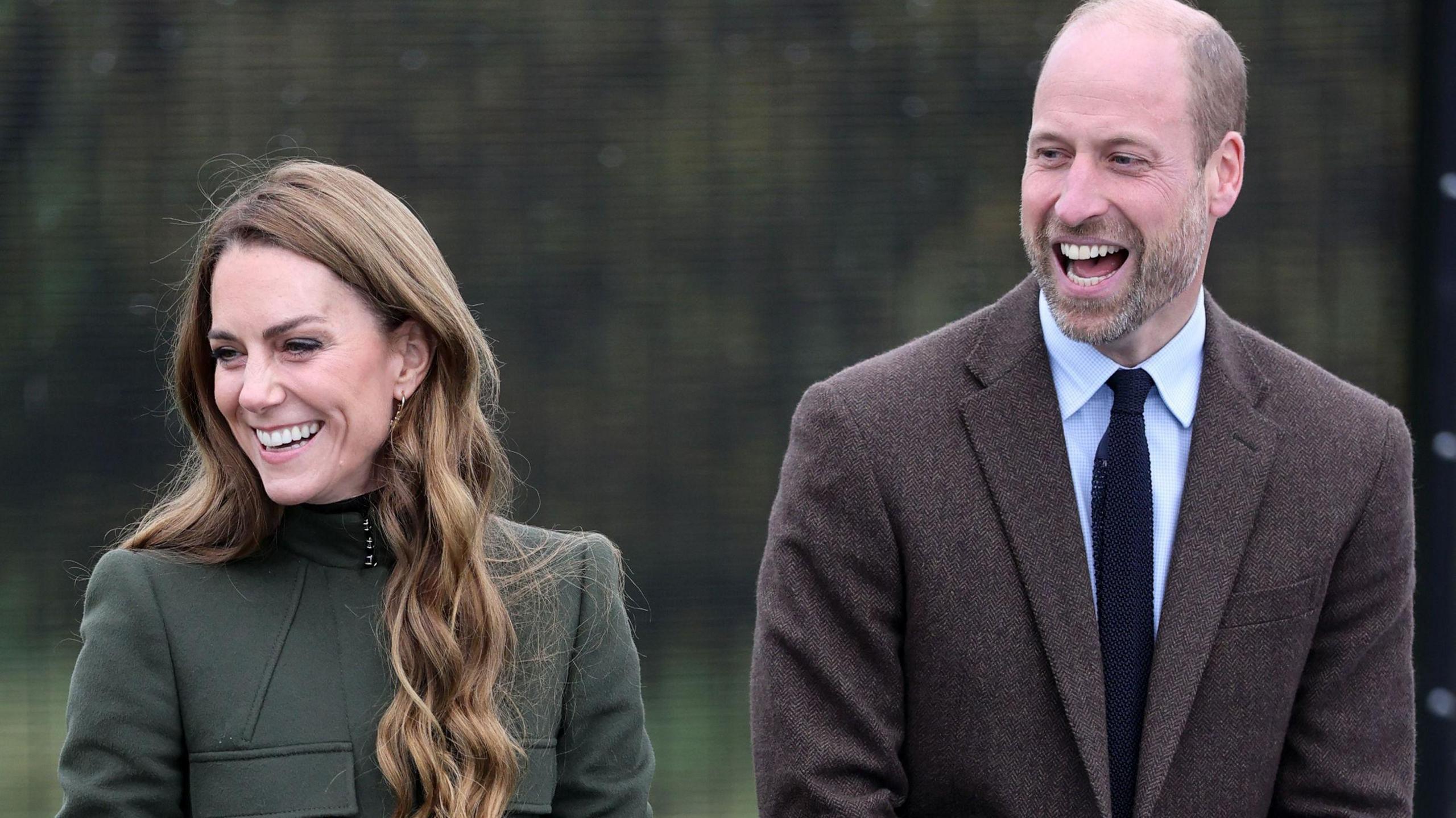 Catherine, Princess of Wales and her husband Prince William smile and laugh during a rope training exercise at the Northern Ireland Fire and Rescue Service (NIFRS) Learning and Development College in County Tyrone. Cathering is wearing a dark olive green coat fastened to the neck and Prince William is wearing a brown blazer, a light coloured shirt and a dark tie.