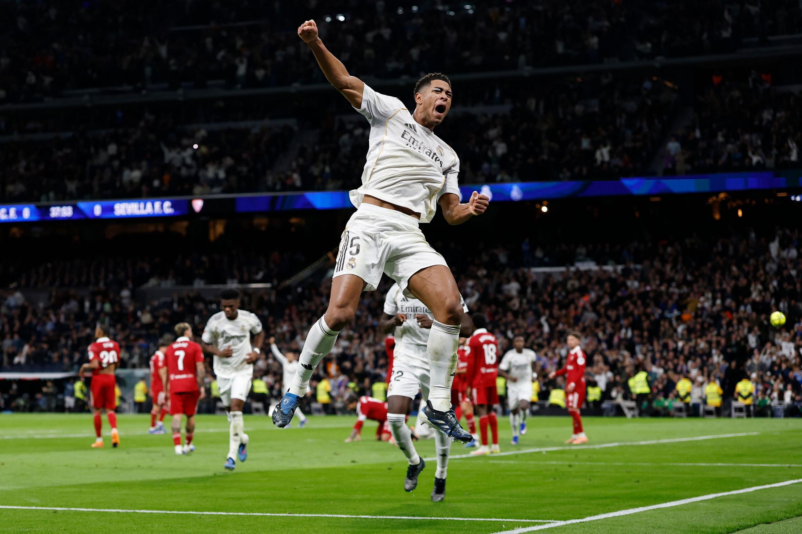 A football player in a white kit leaps into the air with a clenched fist in celebration, surrounded by teammates as they react to a goal. Opponents in red and the stadium crowd fill the background under bright match lighting.