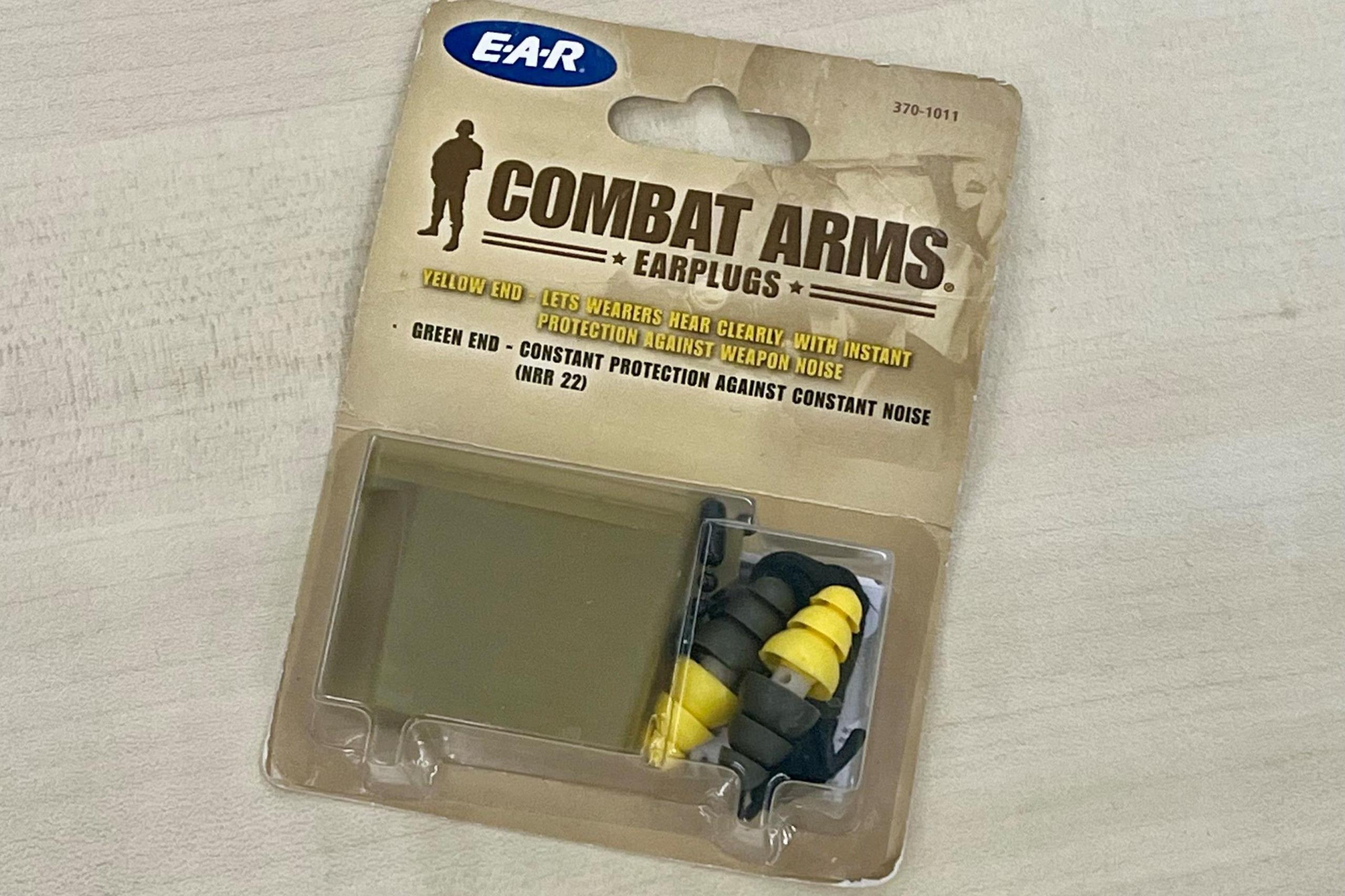 A packet of military earplugs in a box lies on a table. The box says Combat Arms Earplugs, with instructions on how to use them and what they do. A plastic box is in the packaging next to a set of the earplugs, which are yellow on one side and dark green on the other.