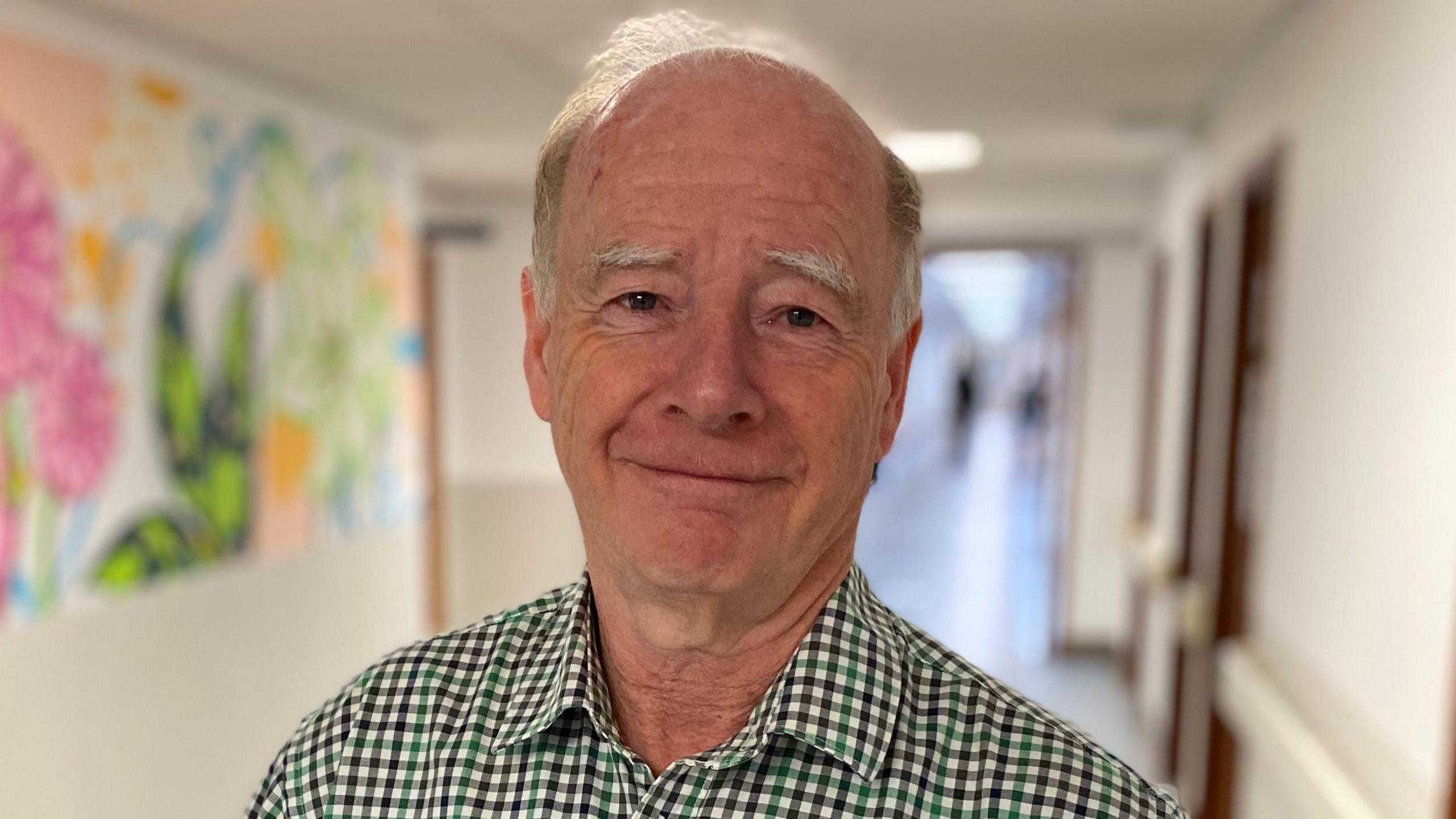 A man called George Oswald looks at the camera while standing in a hallway. He is wearing a green and black gingham shirt. A colourful mural is on the wall over his right shoulder.
