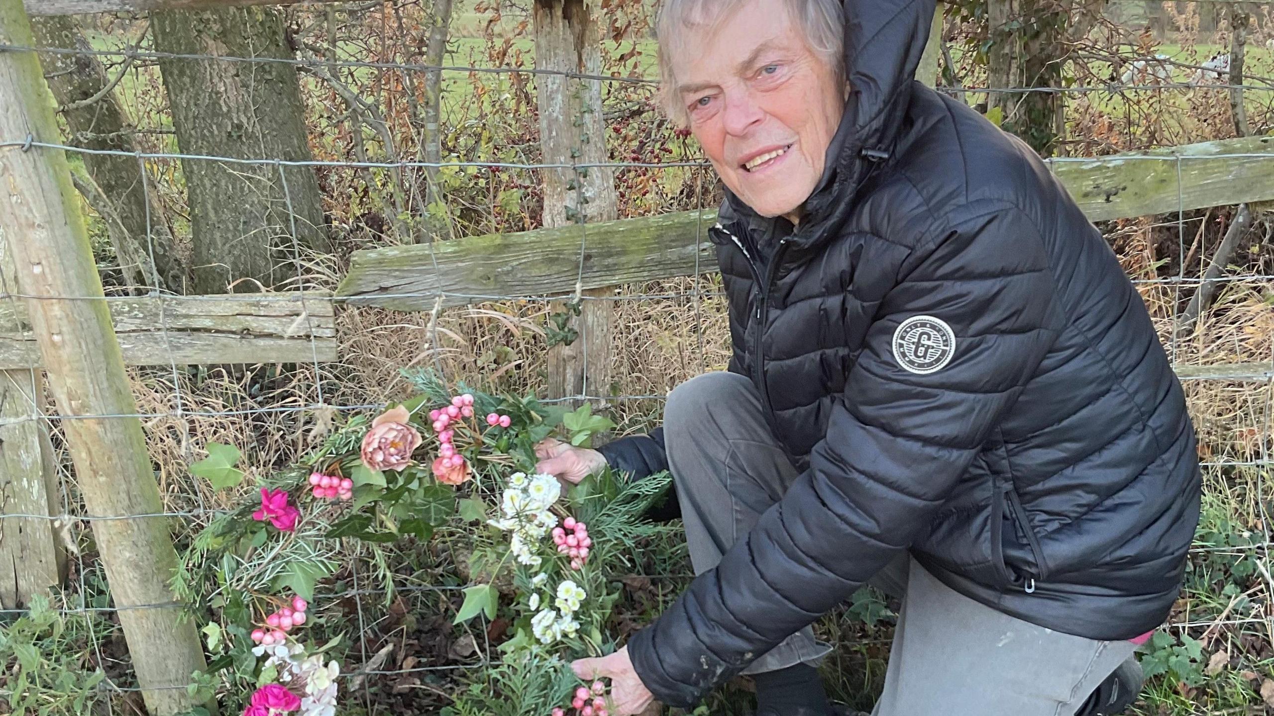 A man in a black puffa jacket and grey joggers lays a floral wreath next to a wooden and wire fence in a grassy field.