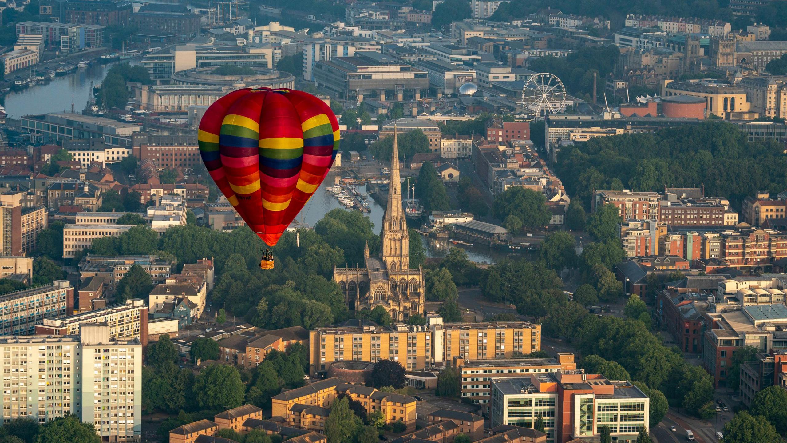 A multi coloured hot air balloon flying over Bristol. The city landscape, consisting of buildings and water, can be seen below.