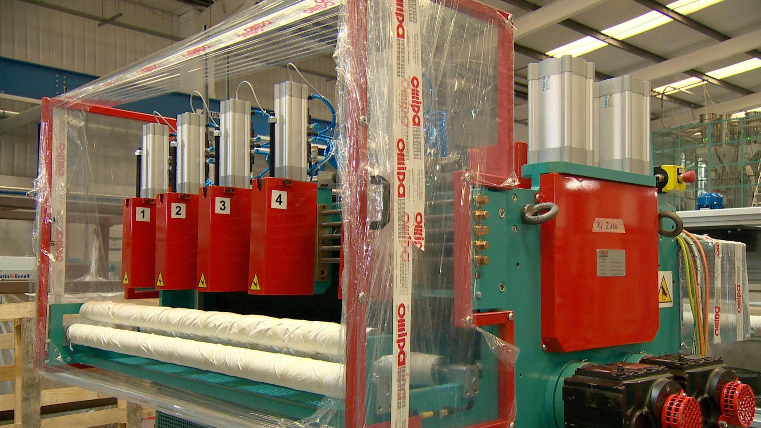 A large turquoise coloured machine, with red numbered cyclinders and two large rollers still wrapped in plastic packaging