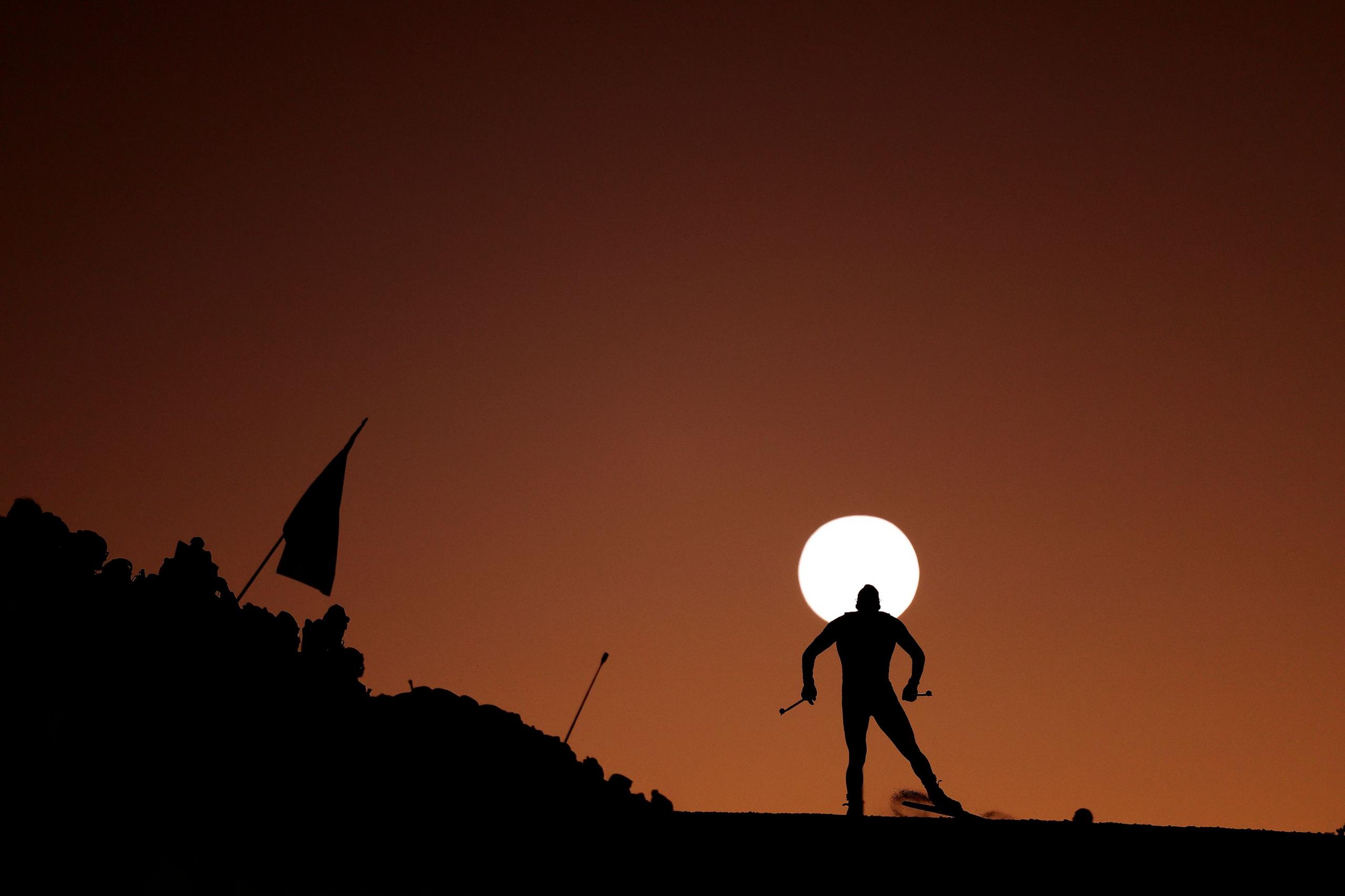 A lone skier is silhouetted against a deep orange sky, gliding along a ridge as the bright sun sits directly behind them. A flag and rocky terrain frame the scene, creating a dramatic sunset backdrop.