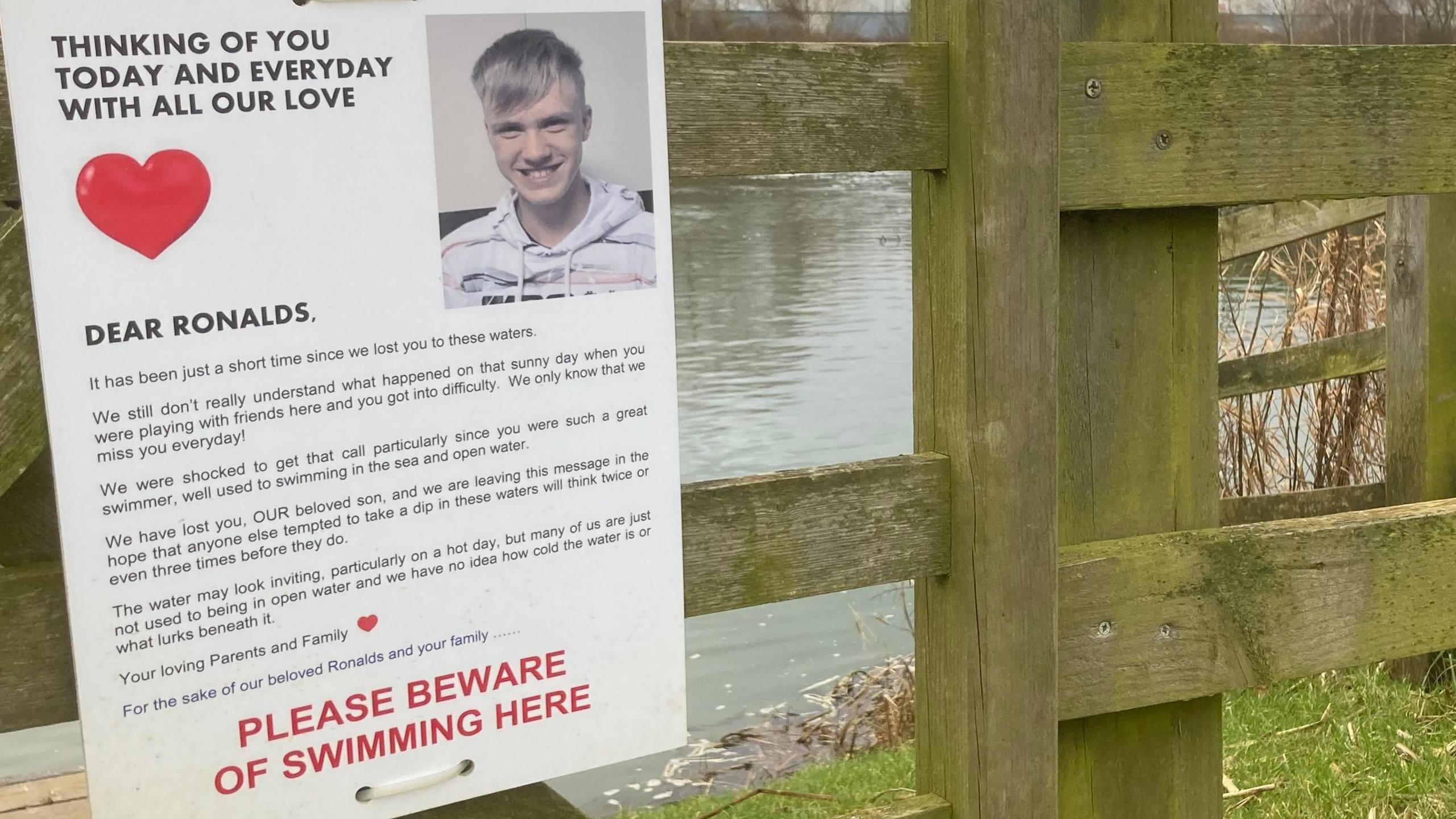 A wooden fence by a river, with a grass bank visible in the foreground. A sheet of card is fixed to the fence with string. It reads "thinking of you today and every day with all our love, dear Ronalds".  Below further text, there is black writing which says "please beware of swimming here".