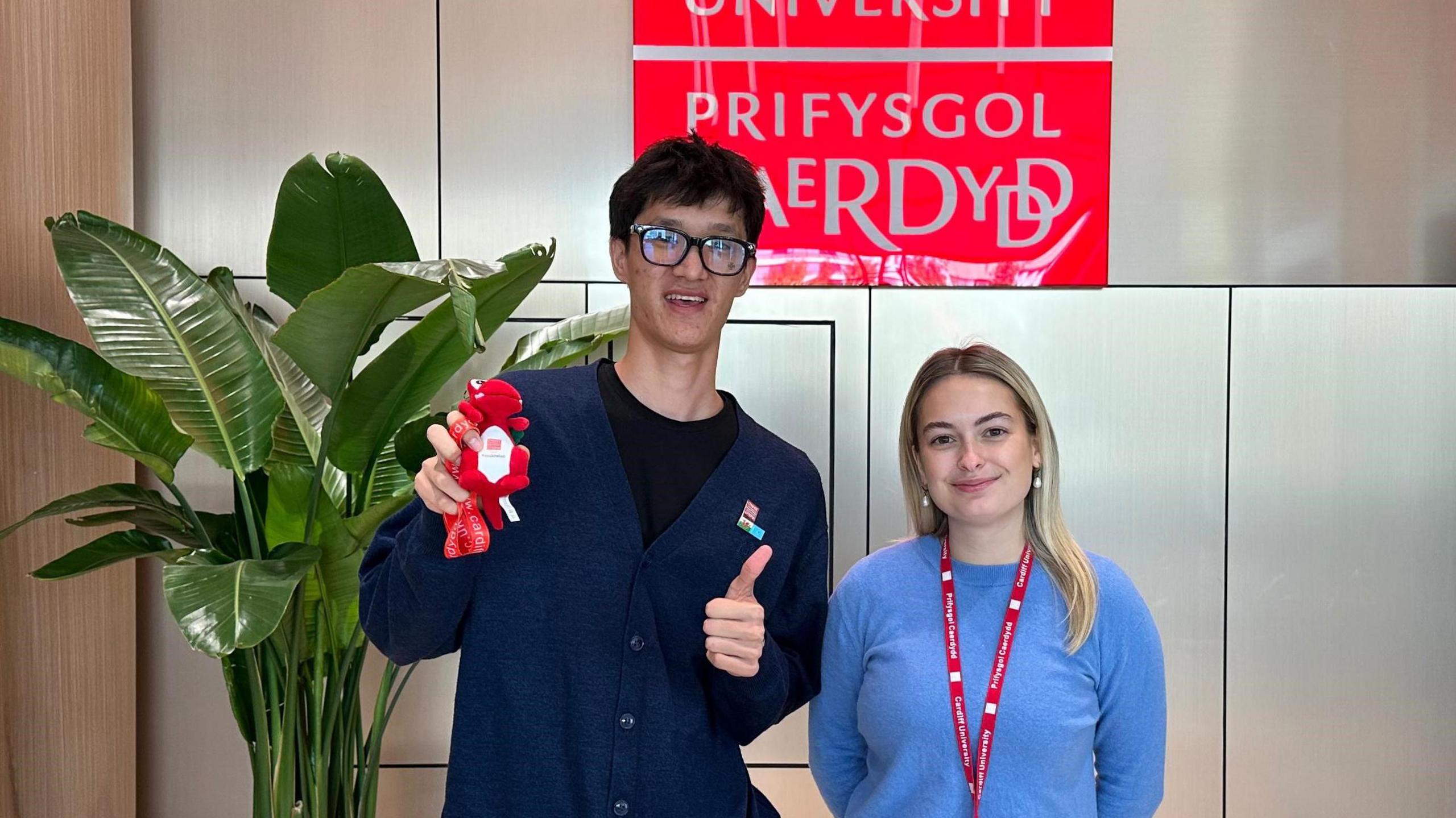 A young man with glasses holding a small red dragon soft toy and with thumbs up, standing next to a young woman with a blue jumper and red lanyard in front of a Cardiff University sign.