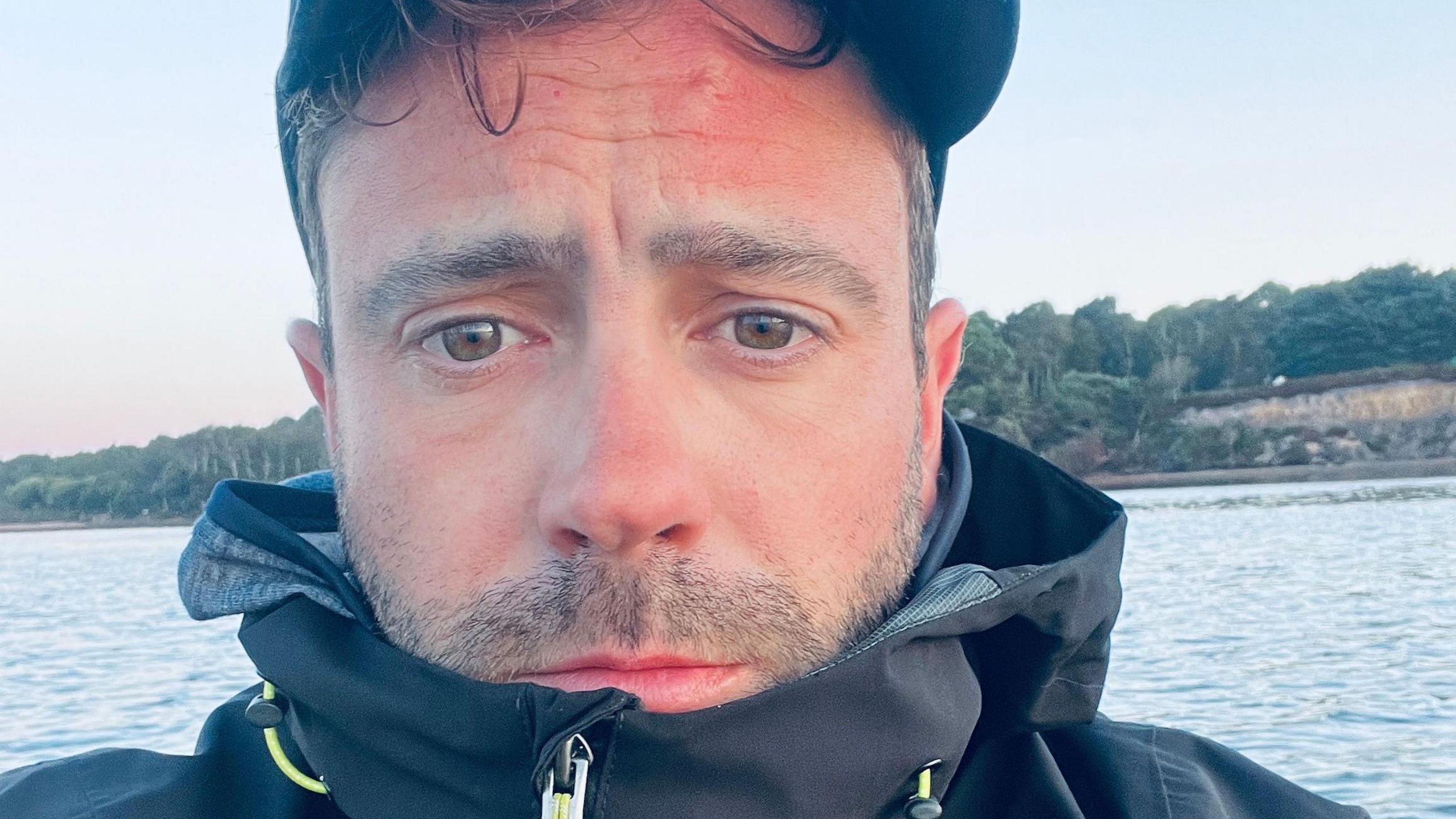 Close-up selfie of David Haze wearing a cap and a coat zipped up over his chin. He is surrounded by water and is looking tired with a furrowed brow