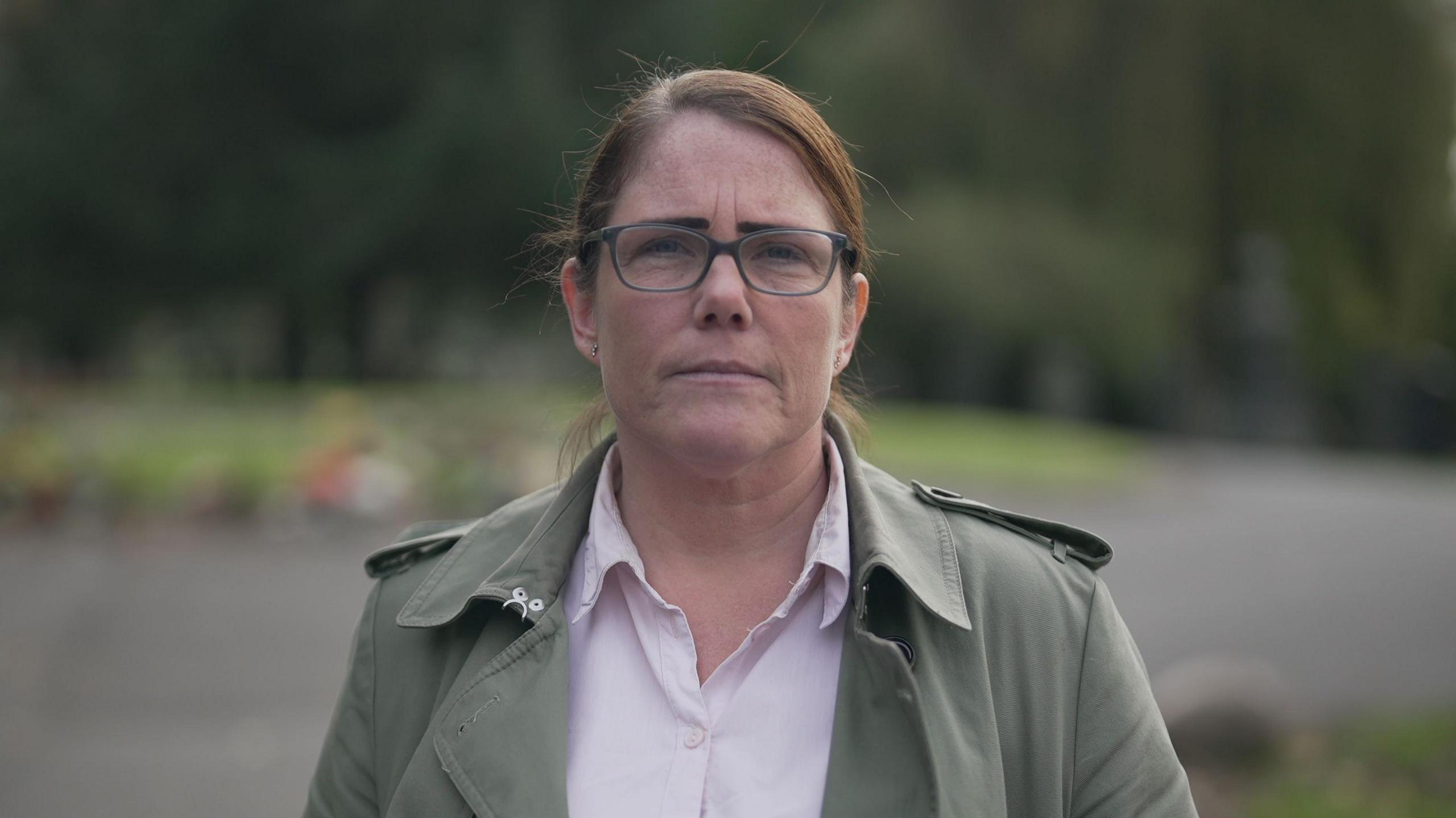 Kate Pollitt, with brown, swept back hair and wearing glasses, is photographed in a park. She wears a light green jacket and an open white shirt.