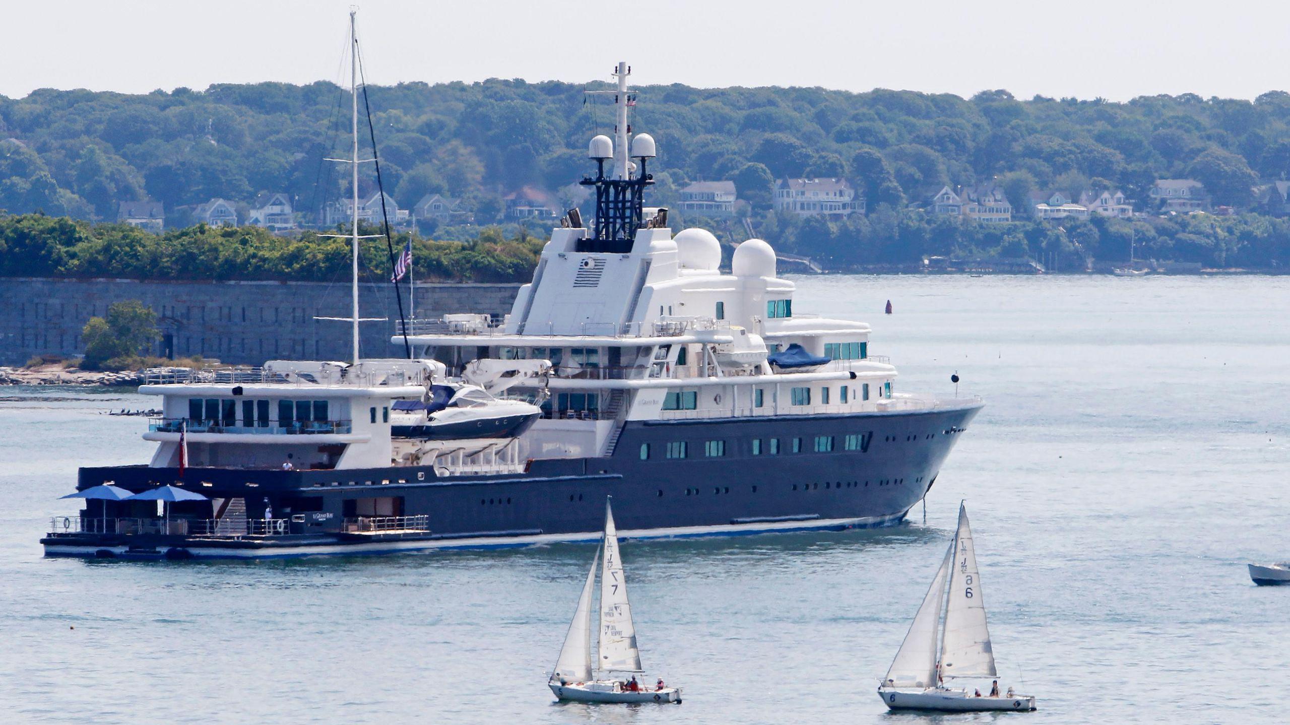 Le Grand Bleu, a large yacht with a navy blue keel and white upper decks, with parasols visible on the large swim platform at the rear, anchored near Portland in Maine, USA, with small sailing boats passing by.