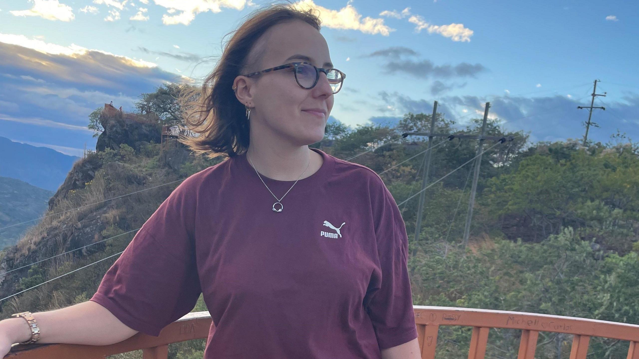 Jesse is seen standing on a platform what overlooks a forest in Honduras. She is glancing away from the camera and is wearing a red t-shirt and glasses. 