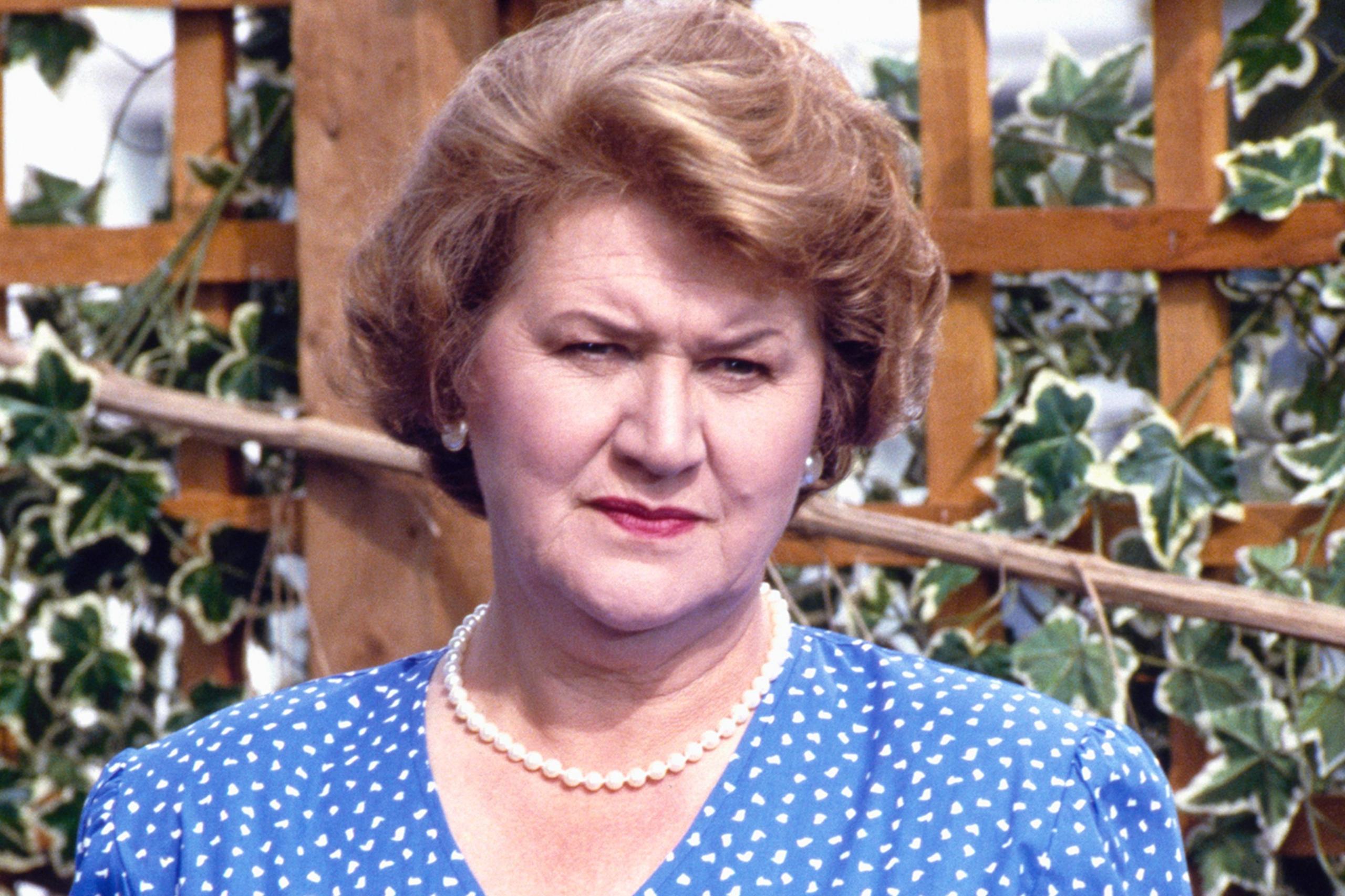 Patricia Routledge ka veshur një fustan blu me një model me pika të bardha dhe një gjerdan me perla të vetme. Sfondi paraqet një rrjetë druri të zbukuruar me gjethe jeshile dredhke, që sugjeron një ambient kopshti ose verande të jashtme. Ndriçimi duket natyral, duke treguar ditën.