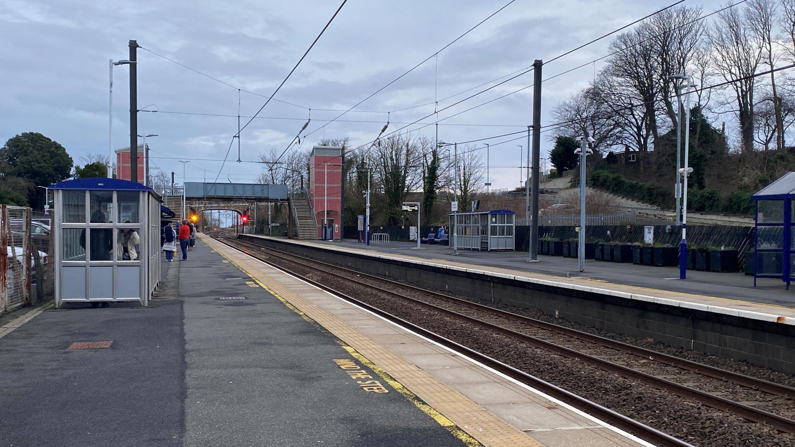 Alnmouth station which has two platforms, one for each direction. In the foreground is a shelter for passengers and further back you can see a footbridge connected by a lift enclosed in a pink tower.