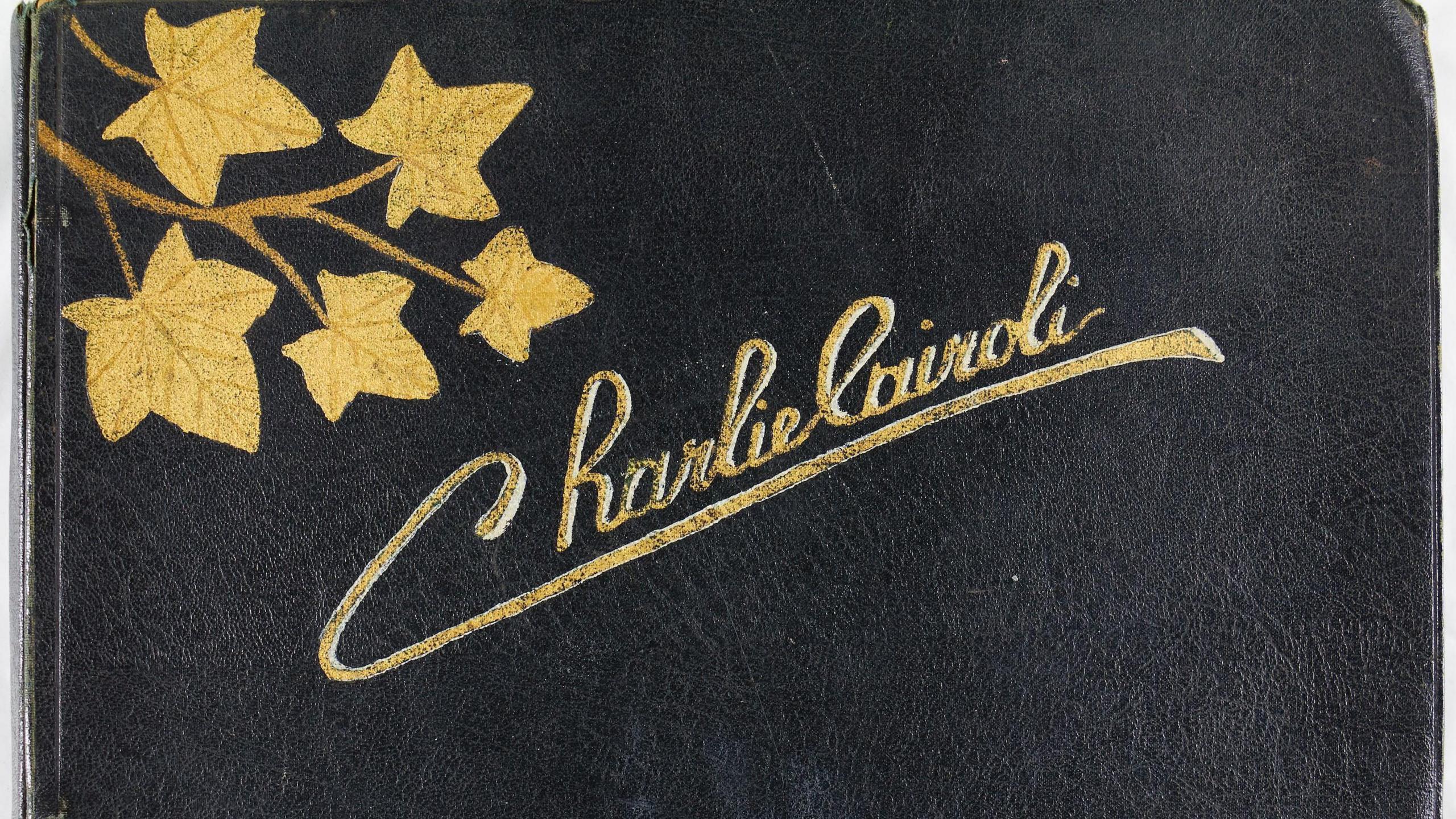 A black leather book with Charlie Cairoli painted on it in gold and leaves painted in the corner in gold.