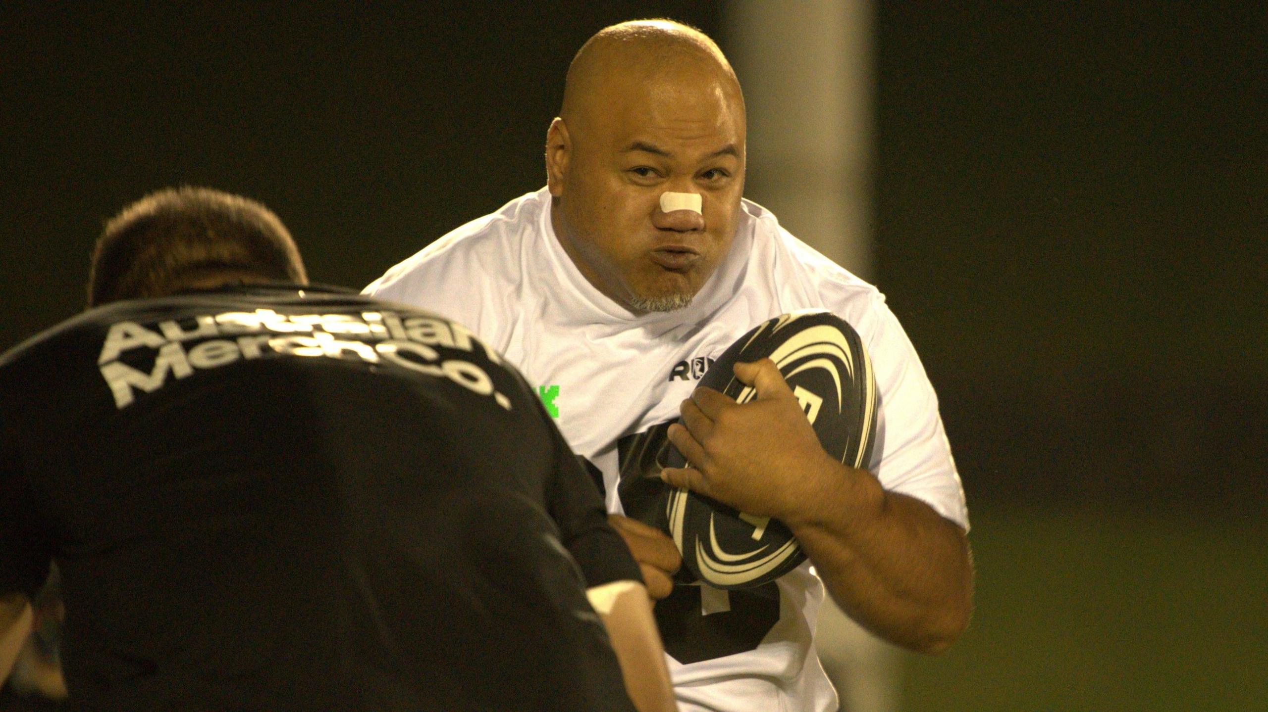 A man wearing a white shirt while holding a rugby ball runs at another man wearing a black shirt who is preparing to tackle him