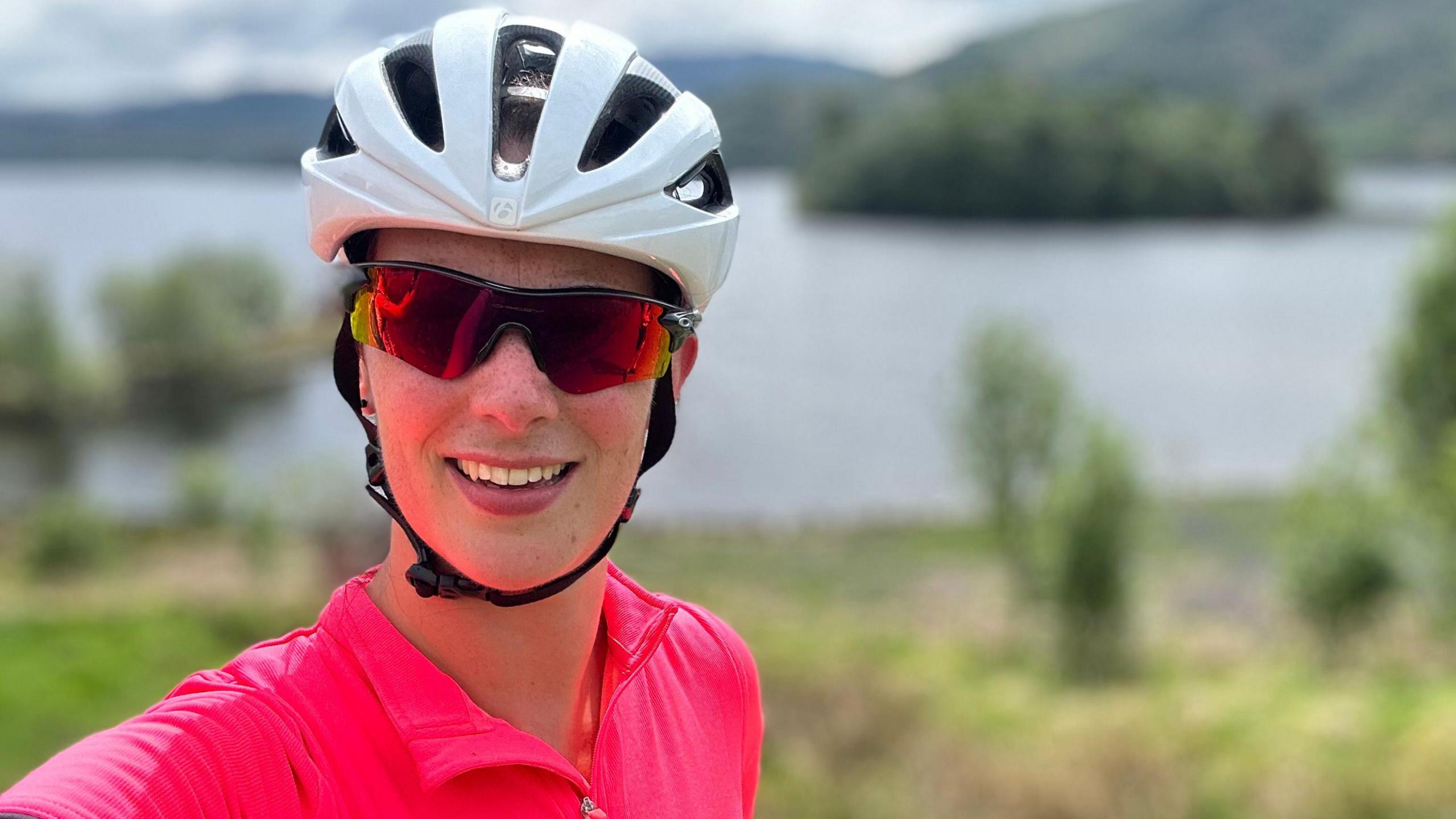 Bex Hind, who is wearing a white cycling helmet, sunglasses and a bright pink top, smiles at the camera. There is a lake in the background and green mountains.