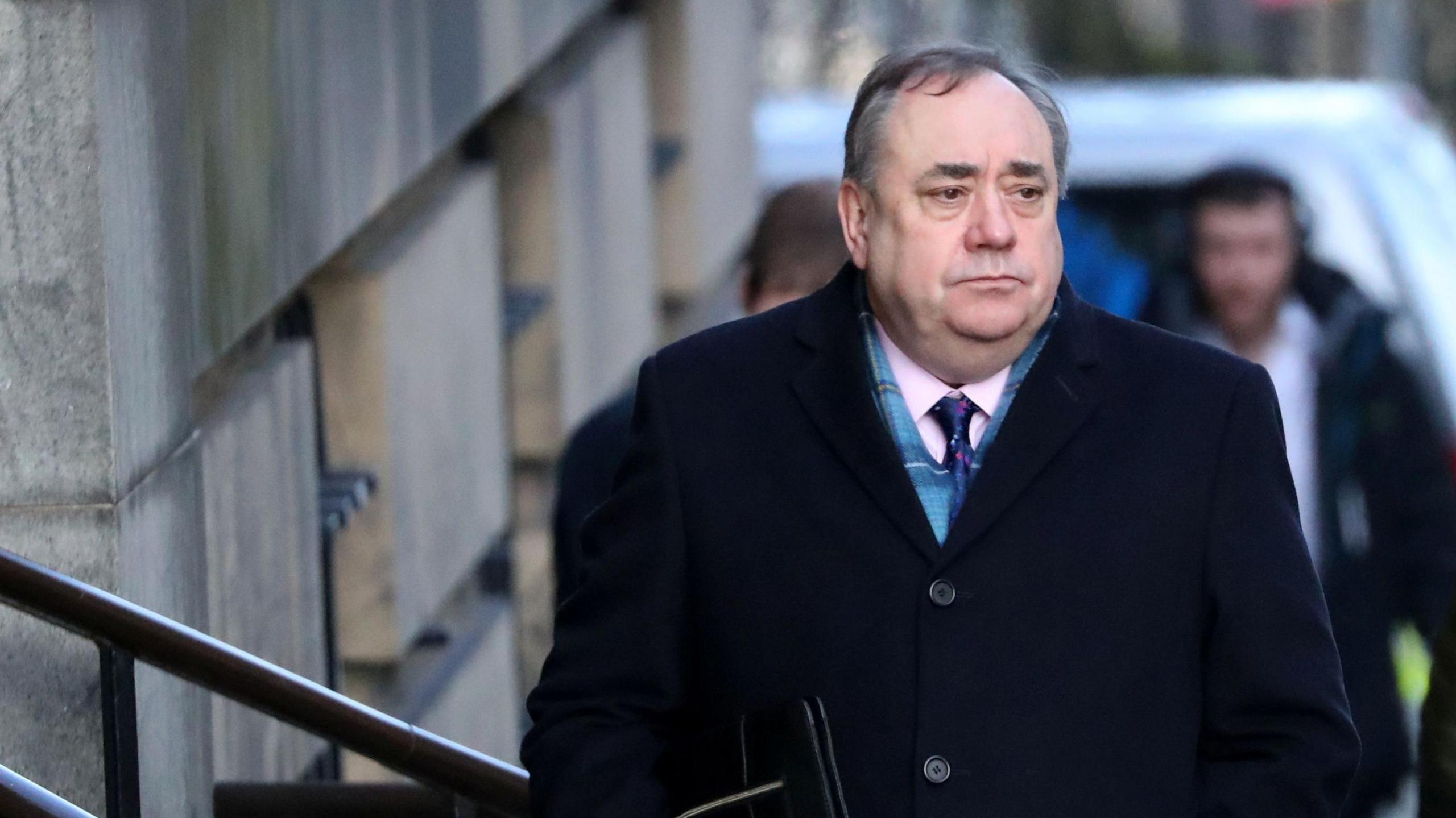 Alex Salmond walking into court in Edinburgh. He is wearing a dark coat, scarf and pink shirt with blue tie. He is looking to his left and appears in a sombre mood. Two men can be seen in the distance behind him.