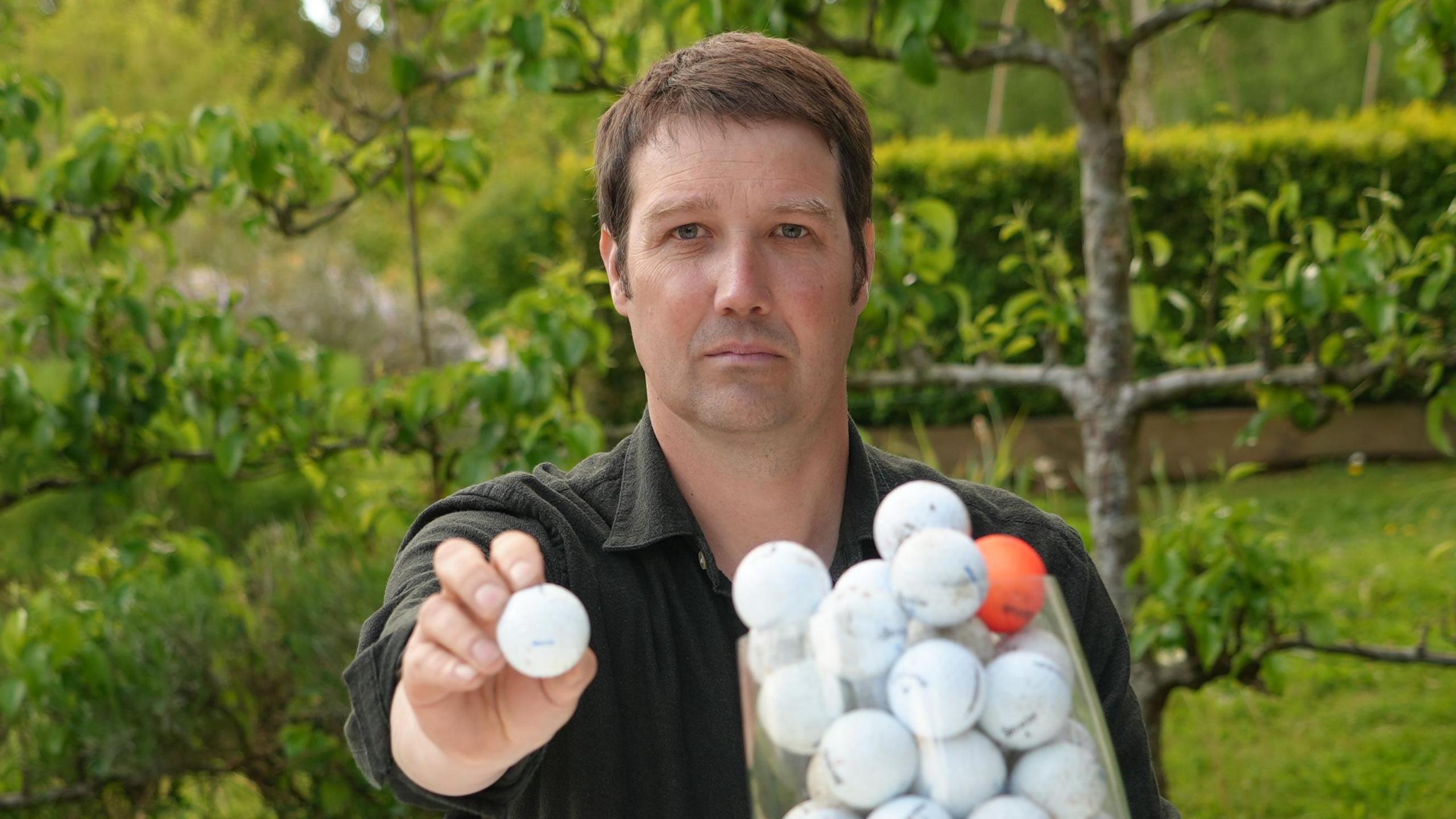 A head and shoulders image of a man wearing a dark coloured shirt while sitting in a garden with trees behind him. He is holding a white golf ball to the camera while looking into the camera. In front of him is a clear receptacle filled with golf balls - all white ones, except for one orange one.