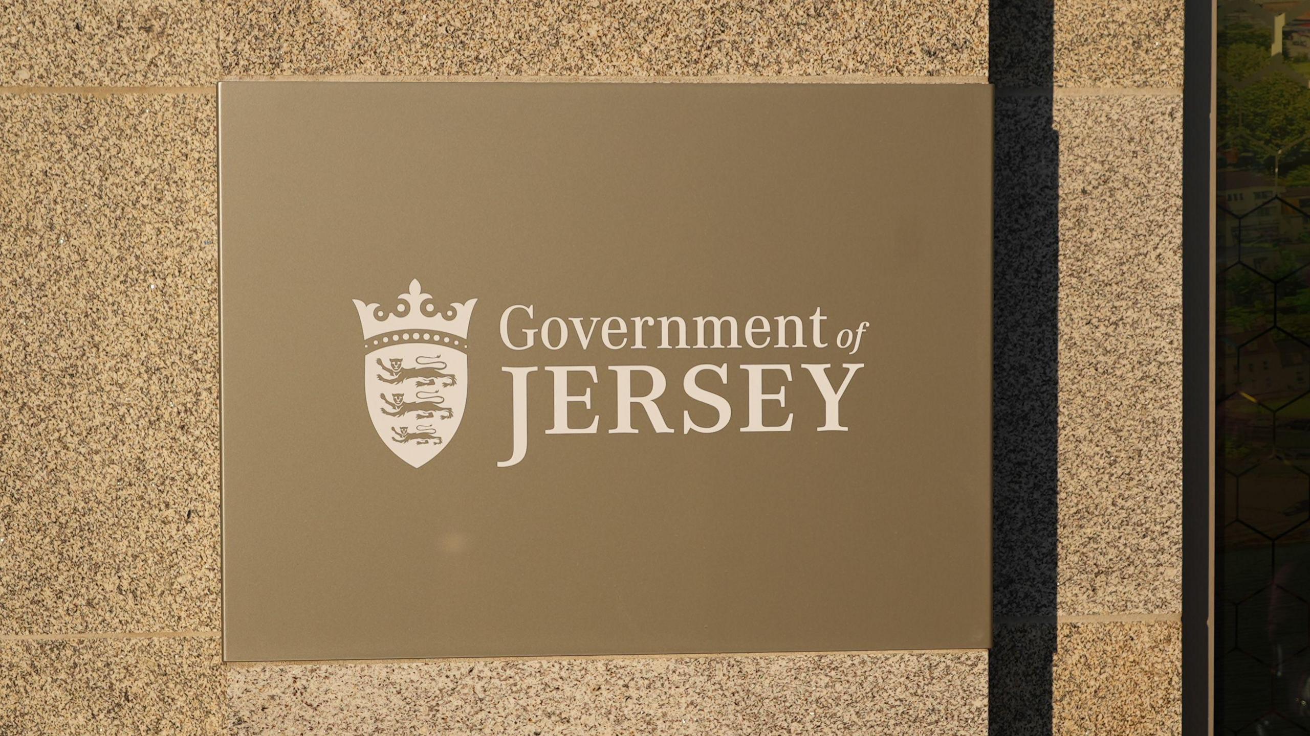 A Government of Jersey sign on its building. The words are white and is on a gold plaque. The wall behind is a light brown.