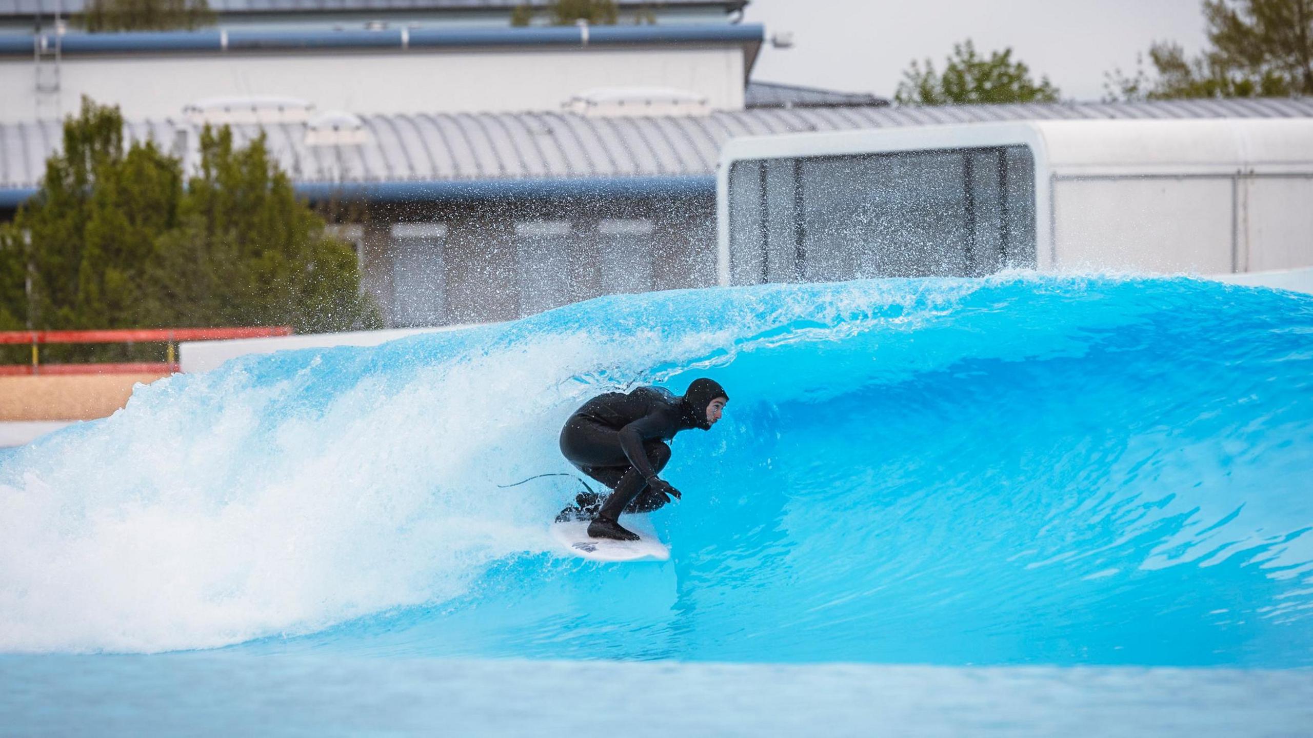 A surfer in a black wetsuit rides a bright blue artificial wave, crouching inside the barrel at an indoor wave pool facility. A modern building is visible in the background.