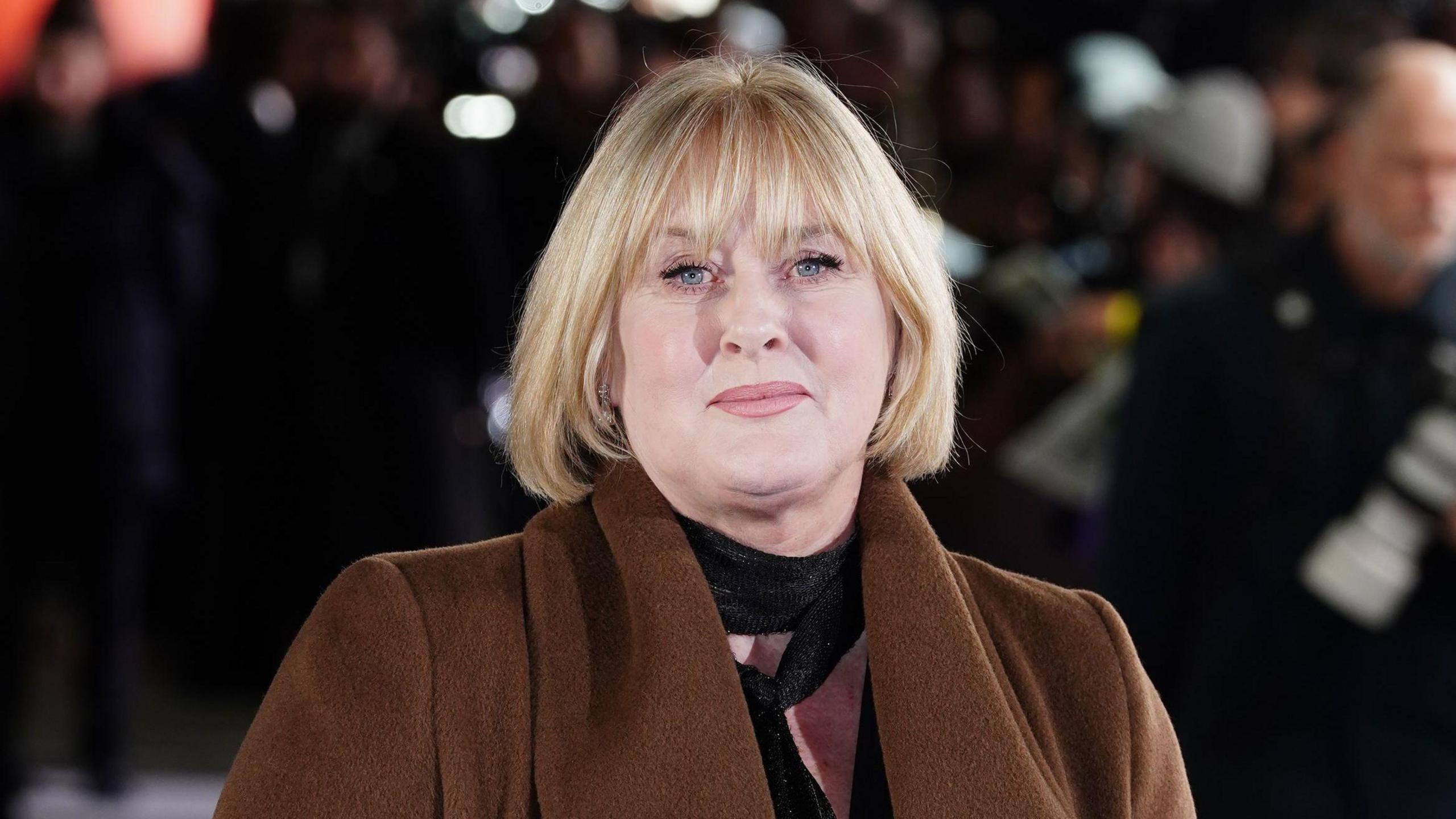 Sarah Lancashire at an outside event. She has blonde hair to her shoulders and is wearing a brown coat.