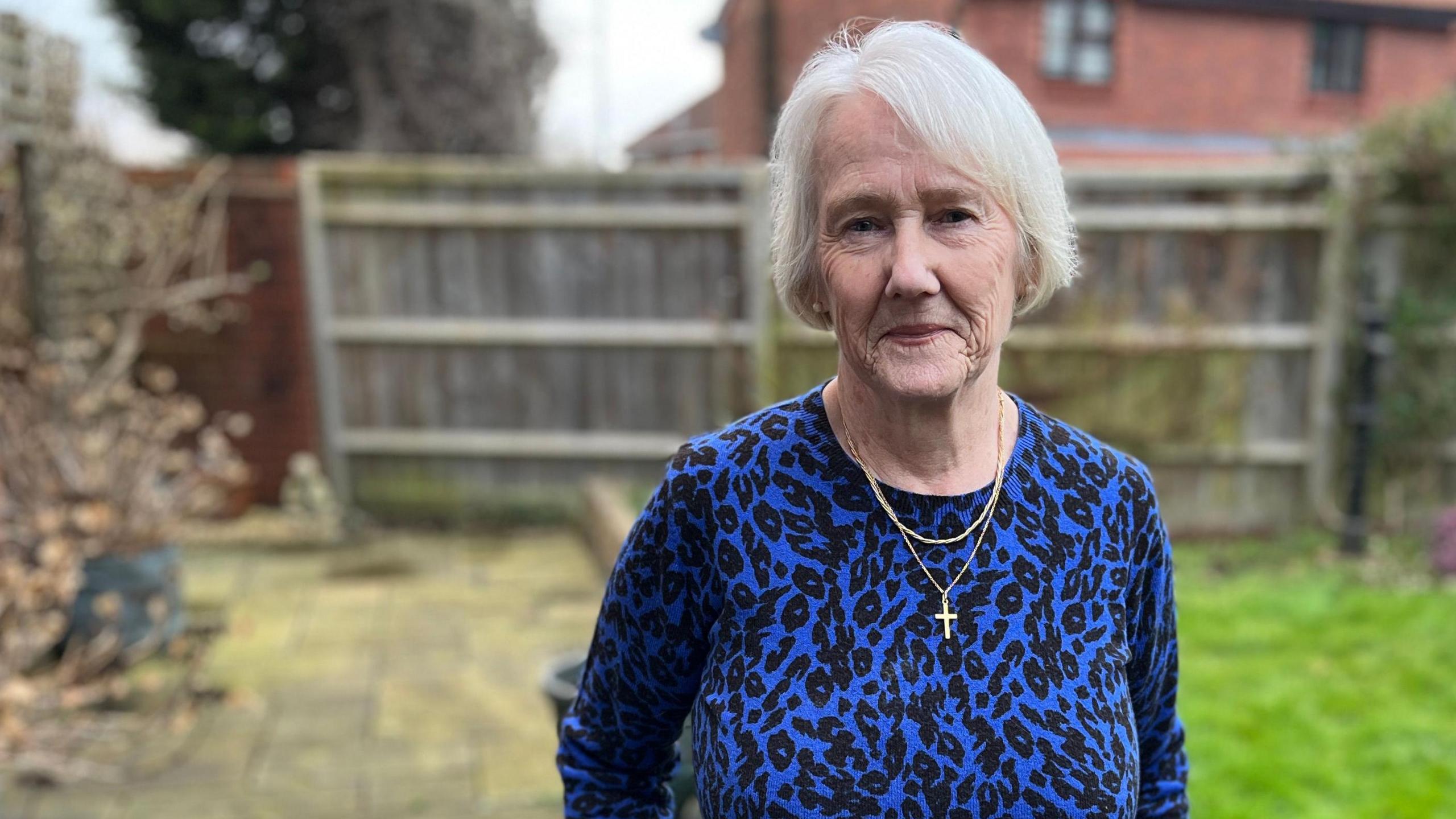 Sue Rowell standing in a garden. She has white hair, wearing a blue and black leopard print top. She is looking into the camera. Behind her is a household garden.