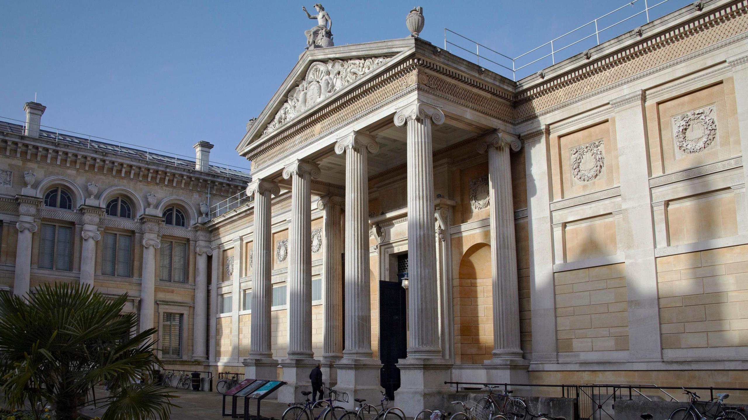 Outside the Ashmolean Museum. There are several bikes at its bike racks. A person is seen approaching the entrance. It is sunny.