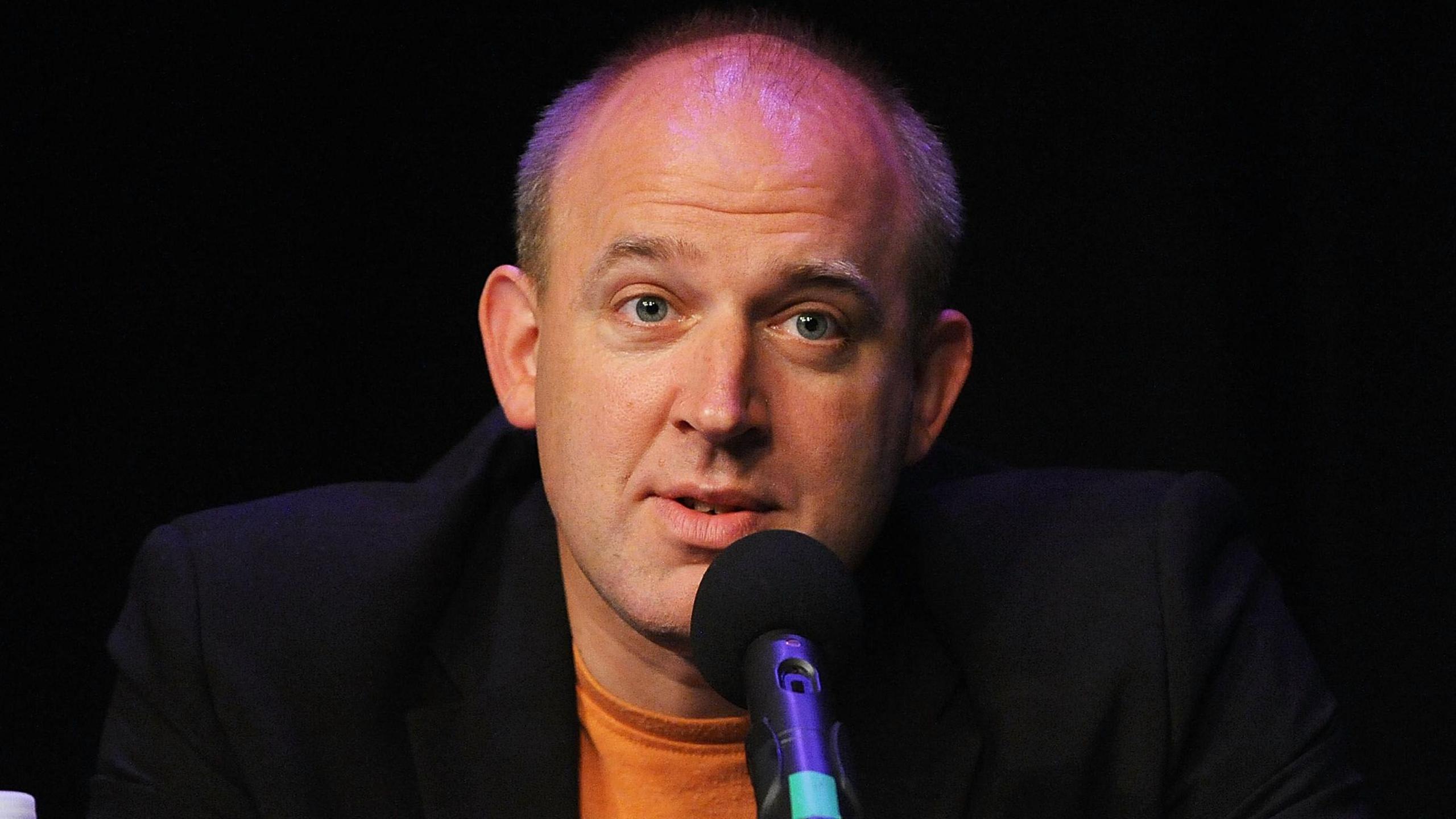 Tim Vine is in the middle of the image sitting in front of a black microphone. He is wearing a black suit jacket and an orange T-shirt.